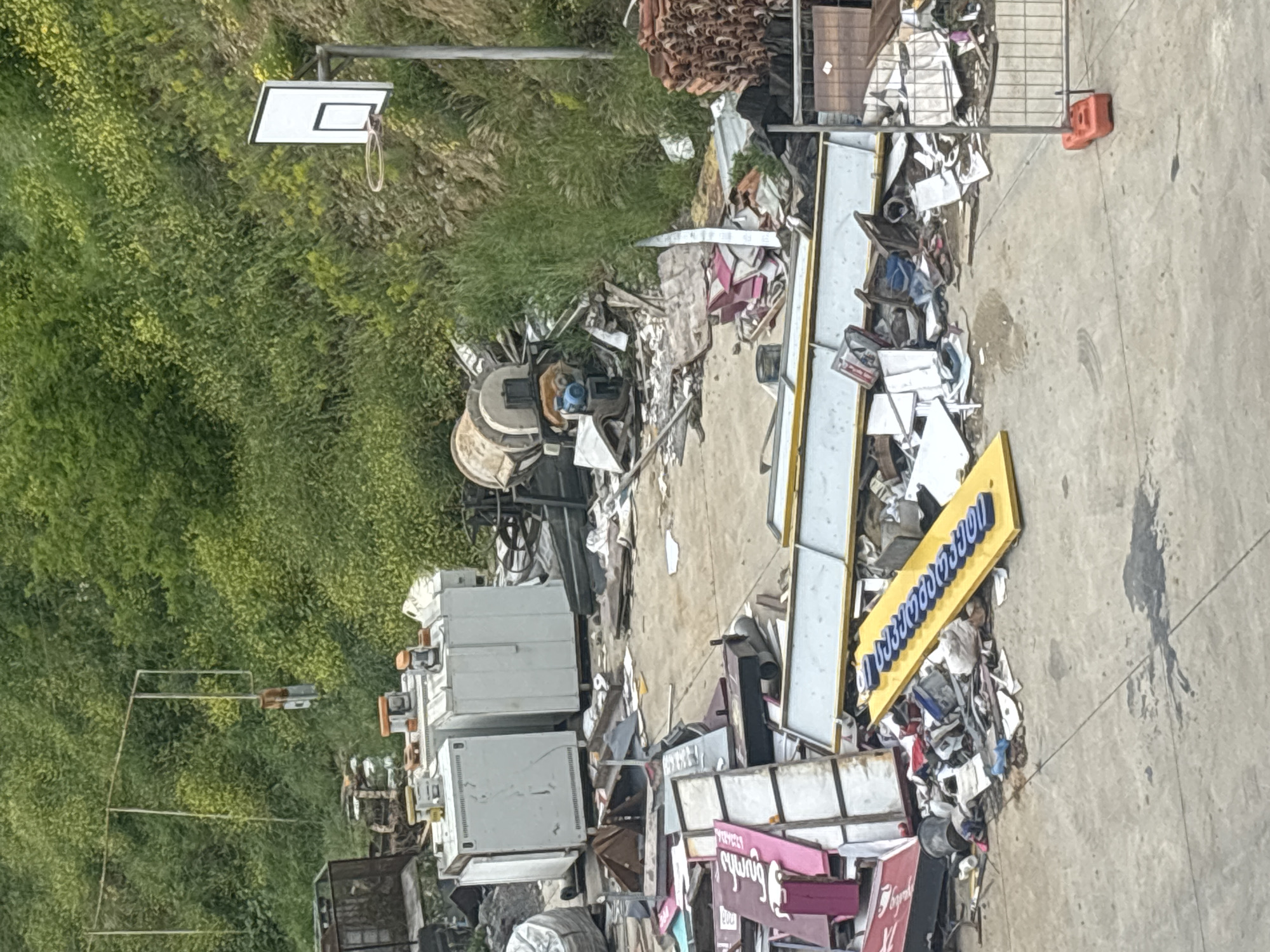 The image shows an outdoor scene, potentially a storage or junkyard, in Tbilisi, Georgia. The area is cluttered with discarded objects including commercial signs, metal scrap, appliances, and debris. A basketball hoop is visible near overgrown vegetation. The ground is paved concrete. No people are apparent.