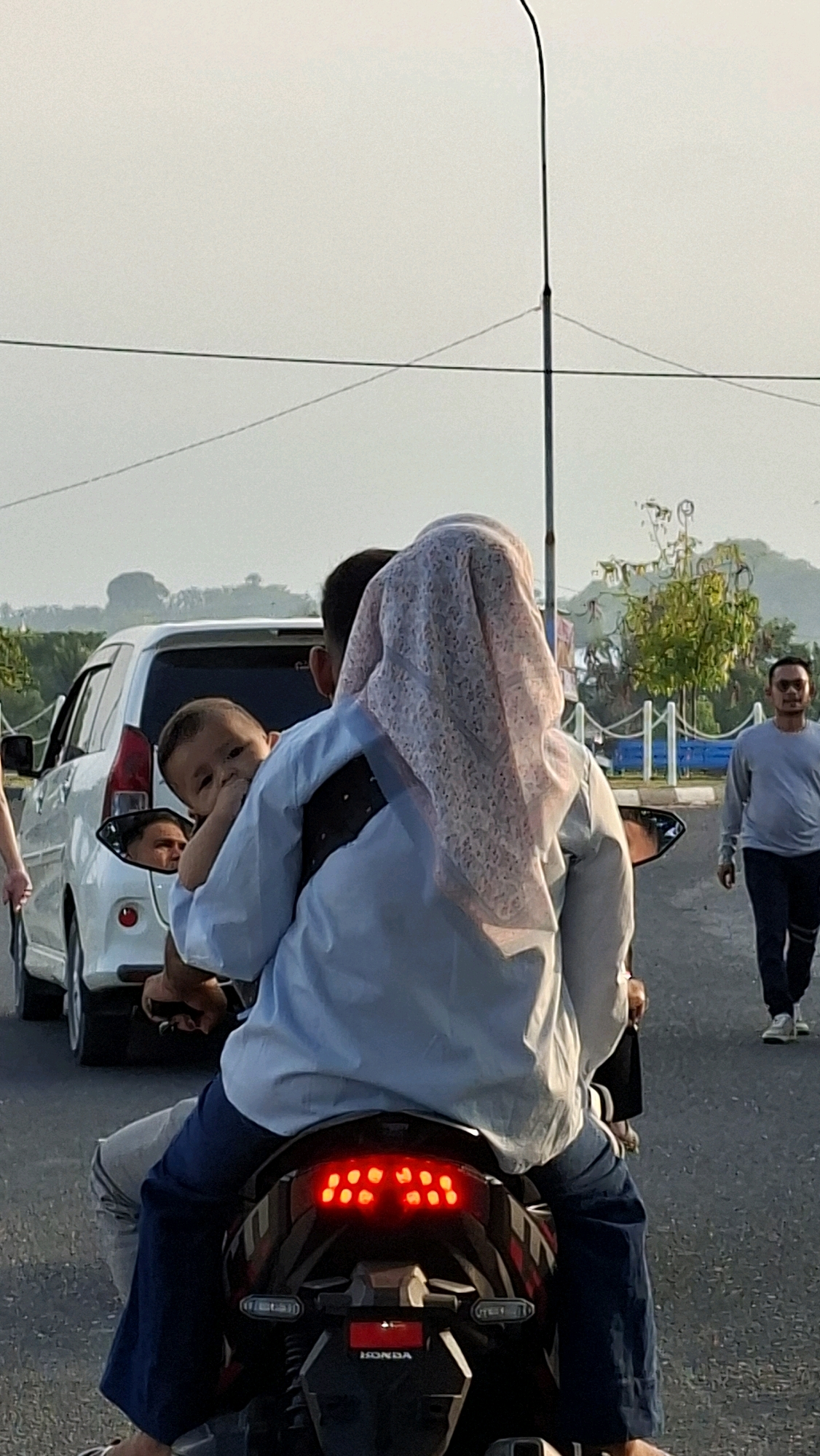 The image captures a scene on a street in Lhokseumawe, Indonesia, likely during daylight hours given the bright sky. The central focus is a person on a motorcycle, holding a young child. The child, looking towards the viewer, is being held by someone wearing a light blue shirt and a patterned headscarf, with a dark vest over the shirt. A white car is visible to the left of the motorcycle. In the background, a man in a grey long-sleeved shirt and dark pants is walking away from the camera. Further in the distance, trees and some distant hills are visible. The street appears to be paved, with some lighting poles and wires overhead. The overall impression is a moment of everyday life captured on a street in the city.