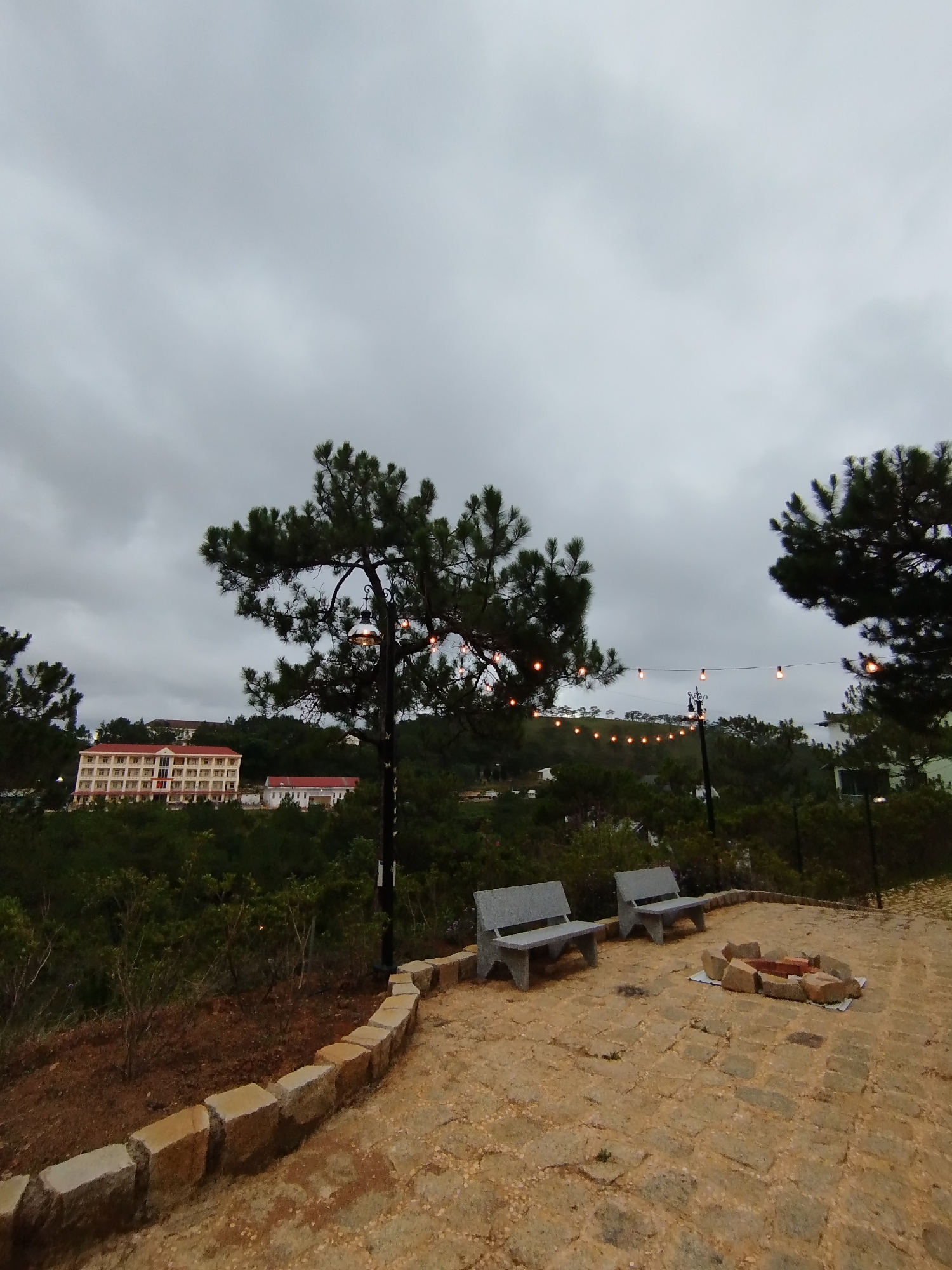 The scene is a paved overlook in Lạc Dương, Vietnam, featuring two gray concrete benches and a circular arrangement of stones, likely a fire pit. The overlook is bordered by a low stone wall and overlooks a valley filled with trees and vegetation. In the distance is a multi-story building with a red roof, along with string lights strung between poles. The sky is overcast. No people are visible in the image.