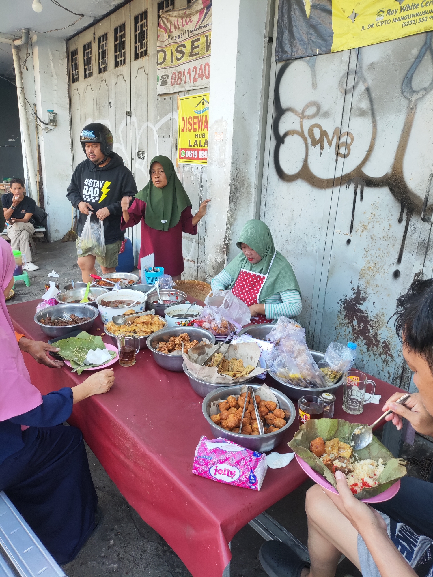 The image captures a street food scene in Cirebon, Indonesia, with a focus on a food stall set up outdoors. A red tablecloth covers a table laden with various dishes served in metal bowls and on banana leaves. These dishes appear to be typical Indonesian fare, including rice, fried items, and possibly some meat dishes. Several individuals are present, including a woman in a green hijab wearing a red and white polka dot apron, seemingly serving food. Another woman in a pink hijab is seated, holding a plate of food. A man wearing a black hoodie and a helmet is standing, holding a plastic bag. A man is also visible sitting in the background, eating. The background shows a building with a closed metal door and some signage, one of which includes the text "Ray White Cirebon" and a street address. Graffiti is visible on the wall behind the stall. The overall impression is that of a busy, informal food service environment on a city street.