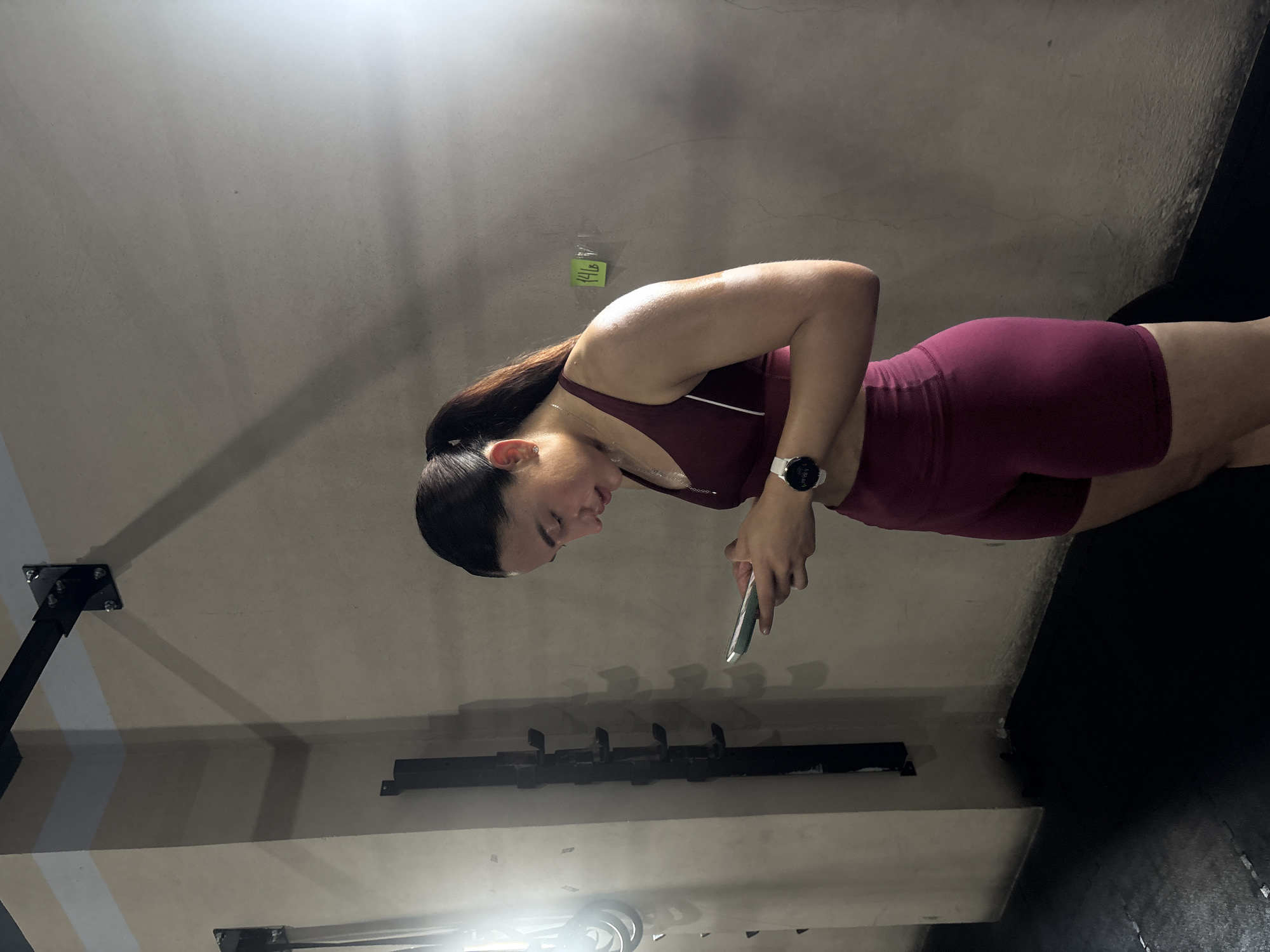 A young woman wearing a maroon workout outfit (sports bra and shorts) is looking at her cell phone in a gym setting. She is standing and holding the phone in front of her. She has dark hair pulled back in a ponytail. The environment includes gym equipment mounted on the wall. The location is the city of San Bernardino Tlaxcalancingo, Mexico.