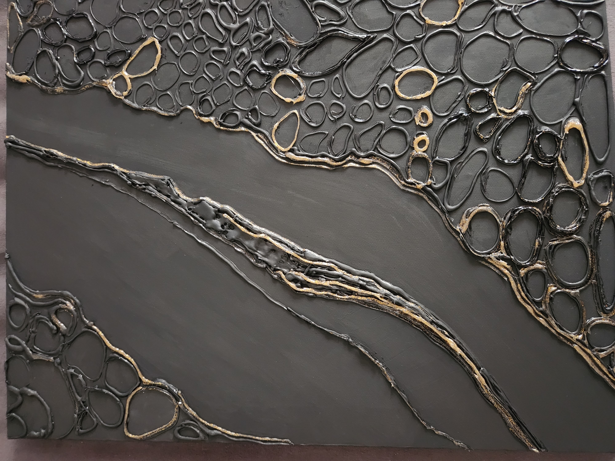 The image displays a close-up, top-down view of an abstract art piece. The artwork is primarily grey with textured elements in black and gold. On the right side, there is a dense cluster of raised, dark, irregular circular and oval shapes outlined in black, with some of these outlines accented by gold. The left side of the artwork features two elongated, wavy textured lines. One line is a thicker, raised black and gold band, and the other is a thinner, more subtle textured line that appears to be a continuation of the pattern from the right. The background is a matte grey canvas. There are no people, actions, interactions, or environmental details related to a city or location like Vniissok, Russia visible in this image.