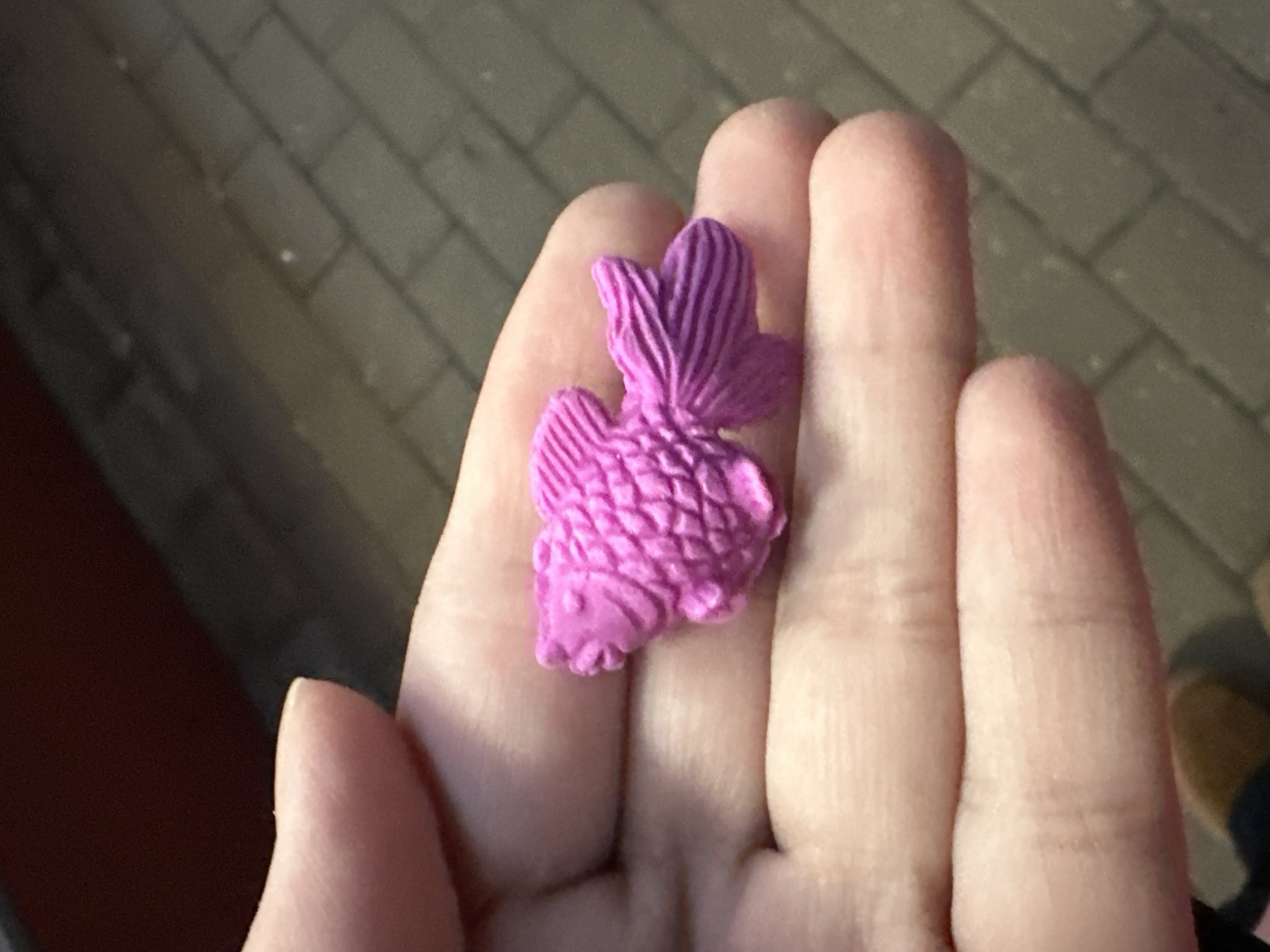 A pink, fish-shaped eraser rests on the palm of a hand. The background is blurred, but shows pavement suggestive of an urban area. The location cannot be determined from this image.