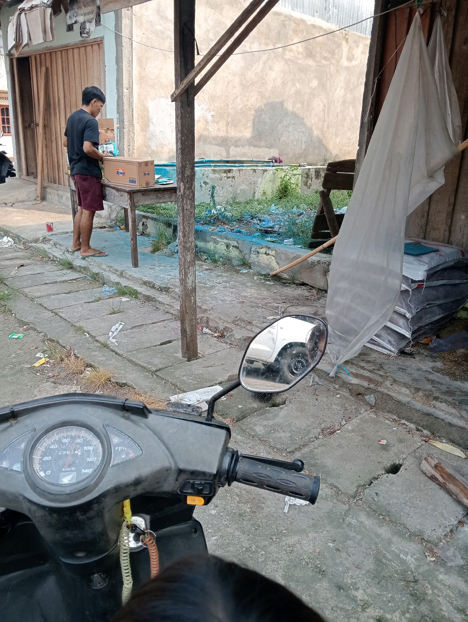 A young man in a black t-shirt and maroon shorts stands on a concrete pathway in front of a wooden building. He is examining a cardboard box on a wooden table. The scene appears to be in a rural or semi-urban area, with a dirt road, scattered debris, and a motorcycle in the foreground. The background shows a textured wall and some greenery. The weather seems to be overcast, judging by the lighting. The location is identified as Pariaman, Indonesia.