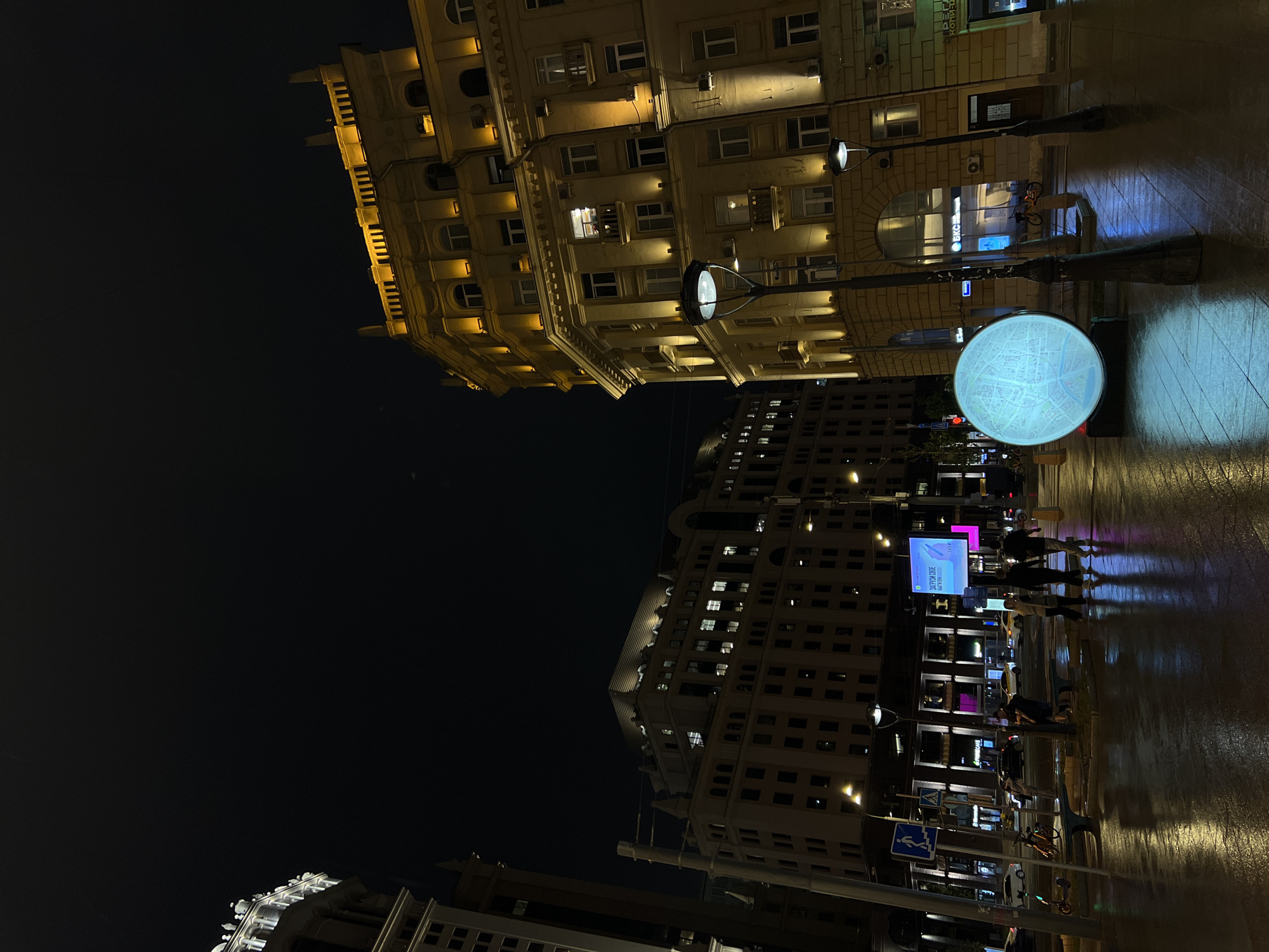 The image is a low-angle shot taken at night in Moscow, Russia. In the upper part of the image, there is a tall, ornate building with many windows, illuminated with a warm yellow light. Below, the street is wet, reflecting the lights of the buildings and signs. There are several illuminated signs. A round light with a map on it stands out. Pedestrians, some blurred due to movement, are walking along the sidewalk. The sky is dark, contrasting with the bright lights of the city. In the lower part of the image there are more buildings in the distance.