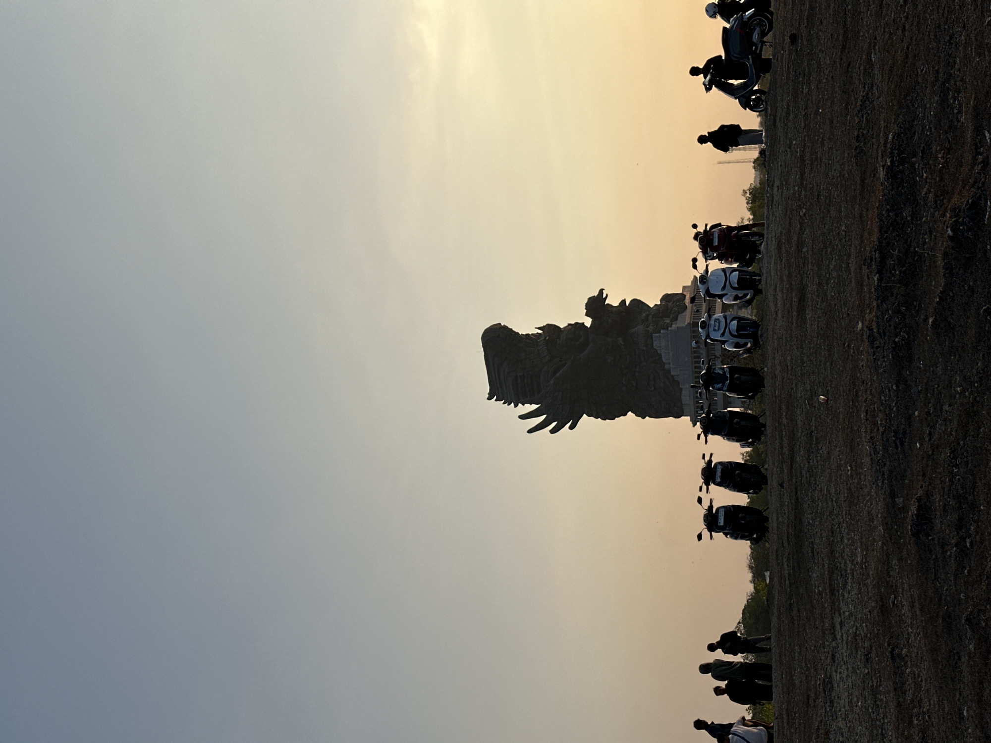 This image depicts a silhouette of a large Garuda statue, a mythical bird from Indonesian folklore, against a gradient sky ranging from light blue at the top to pale orange at the horizon. The statue is the focal point of the scene, situated on a hilltop. In the foreground, a group of people are standing and a line of motorcycles is parked. The ground appears to be dry and barren, with sparse vegetation. The lighting suggests it is either sunrise or sunset. The Garuda Wisnu Kencana Cultural Park in Kuta Selatan, Indonesia, is a likely location for this statue and scene.