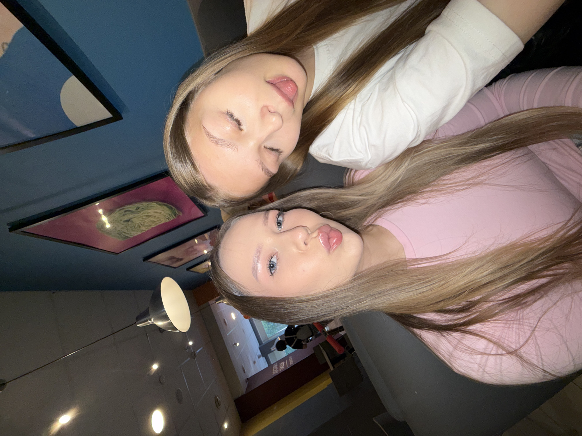 The image depicts two young women taking a selfie in what appears to be a modern cafe or lounge in the city of Chaikovsky, Russia. The woman on the left, with long, straight light brown hair, blue eyes, and wearing a light pink long-sleeved top, is posing with pursed, glossy lips towards the camera. The woman on the right, also with long, straight light brown hair and wearing a white long-sleeved top, is leaning her head slightly, with her eyes closed and her tongue playfully extended.
They are seated on dark upholstered furniture. The ceiling above is grey, featuring several recessed lights and a prominent silver pendant lamp hanging from a wire. The wall to the right of the women is painted a distinct dark teal or blue, displaying two framed art pieces: one with a vibrant pink background and a green, circular, fibrous design, and another with a simple white semicircle against a light blue background. To the left, a dark reddish-brown column or partition is visible, bearing partially obscured Cyrillic text that reads "ДОСТАВКА ПИЦЦЫ" (Pizza Delivery), indicating a Russian-speaking environment. In the background, beyond the women, a doorway or corridor leads to an area where a silhouetted individual, wearing a dark top and a backpack, is seen walking away, carrying a red cylindrical object. Natural light from an exterior window or glass door is visible in the far background.
