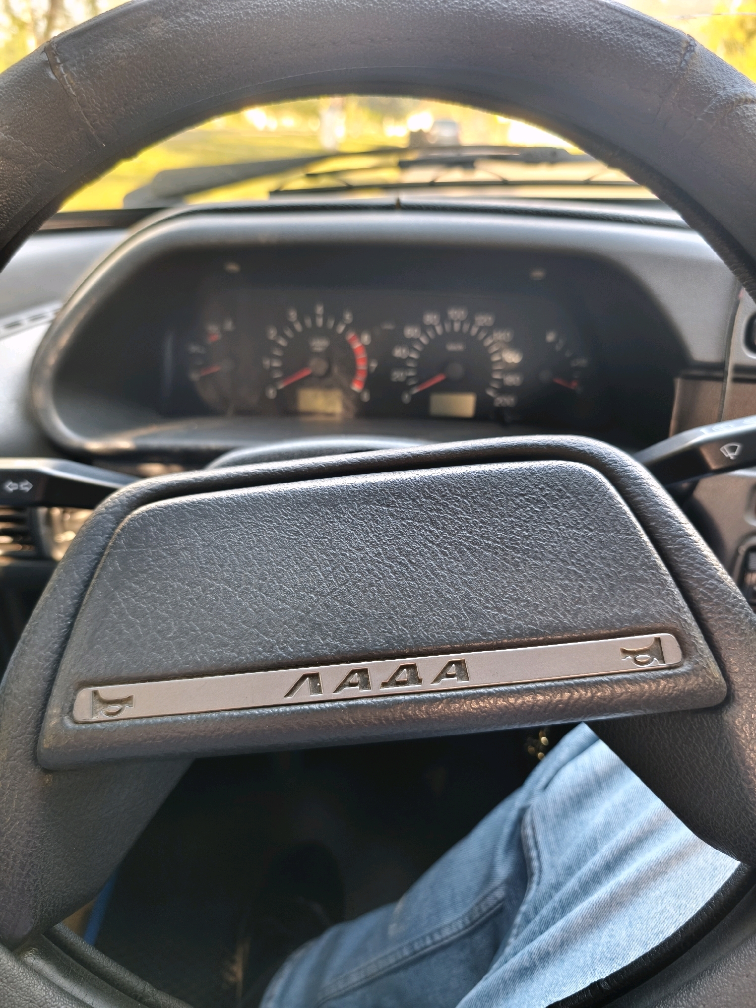The image is a close-up shot from inside a Lada car, likely in the city of Karachayevsk, Russia. The foreground is dominated by the steering wheel and a portion of the dashboard. The steering wheel is black and appears to be covered with a textured material. The dashboard is dark gray, and prominently displays the Lada logo with a horn symbol on either side. Behind the steering wheel, the car's instrument cluster is visible, showing various gauges, including a speedometer. Sunlight streams in from outside, illuminating the dashboard and creating a bright, yellow hue in the background, suggesting it is daytime. The image is framed from the perspective of someone sitting in the driver's seat. The lower part of the image shows the driver's blue jeans. There are no other people or animals visible in the frame.
