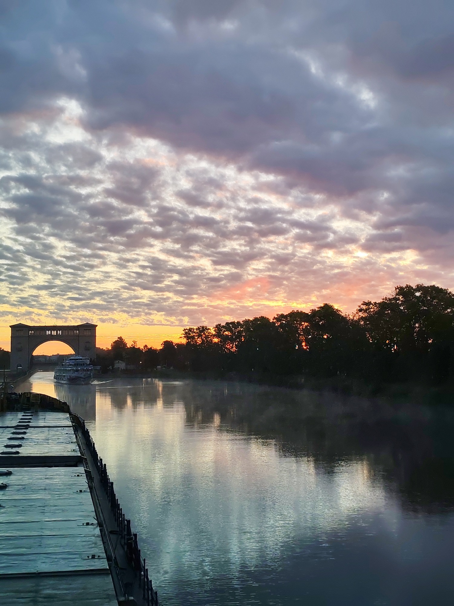 An early morning view of the Volga River in Chelganovo, Russia, shows a barge in the foreground, its deck made of weathered metal planks and equipped with a railing. The river, shrouded in mist, reflects the dramatic sky above. In the middle ground, a passenger ship navigates the waterway, approaching a large arched aqueduct. The aqueduct, constructed of brick, spans the river and serves as a crossing. Silhouetted trees line the riverbank, and the sky is a canvas of clouds painted with the soft hues of sunrise, ranging from soft grays to vibrant oranges and pinks.