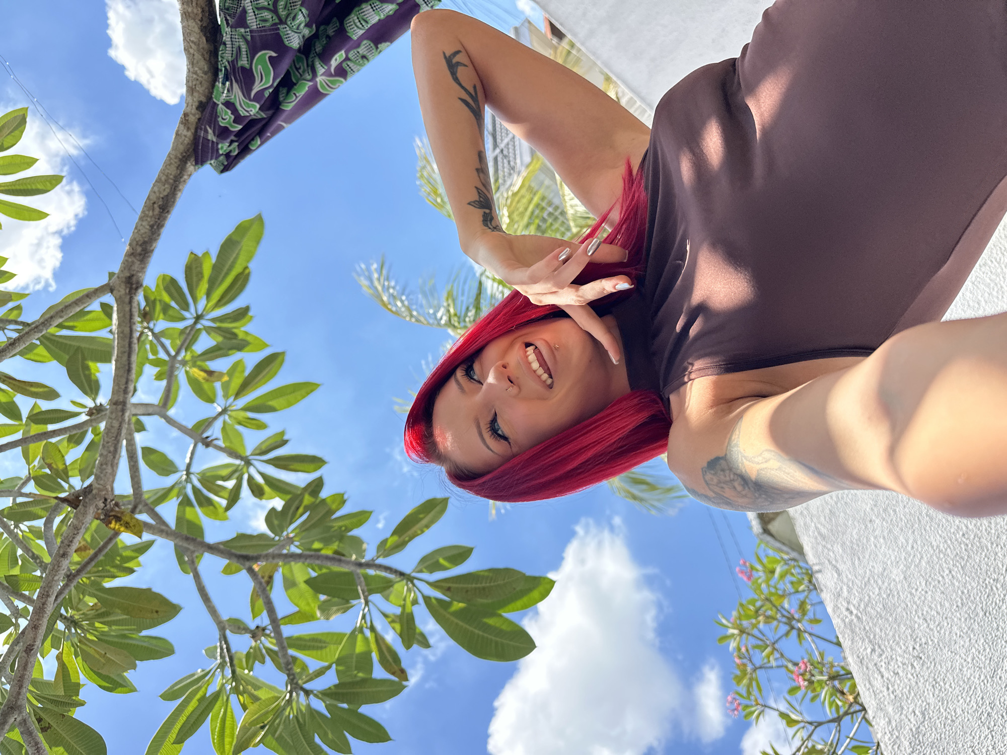 The image is taken from a low angle, looking up at a woman with bright red hair. She is smiling and posing with her hand near her face. Her fingernails are silver. She has tattoos on both arms. She wears a brown dress. The background features a blue sky with scattered clouds, green foliage, and parts of a white building. A piece of green and black patterned fabric hangs from a branch in the upper left corner. Based on the presence of tropical foliage and the style of architecture, the location could be in North Kuta, Indonesia.