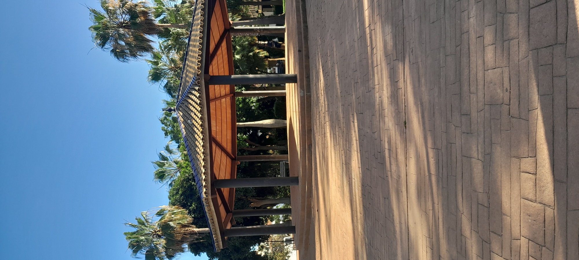 The image captures an outdoor scene under a brilliant, cloudless blue sky, characteristic of Manilva, Spain. On the left, a prominent covered walkway or pergola extends horizontally across the frame. This structure is supported by several dark, sturdy columns and features a distinctive tiled roof. The upper part of the roof showcases a pattern of iridescent blue-green and beige-colored curved tiles, while the underside of the canopy consists of light brown, possibly wooden, panels.
Numerous tall palm trees with green fronds are visible around and behind the walkway, enhancing the warm, tropical atmosphere. Through the columns and foliage, a light-colored, elongated, and somewhat abstract sculpture or decorative element can be seen in the mid-background.
The right side of the image is dominated by a wide paved pathway, made of a reddish-brown or light brown textured material, resembling stamped concrete designed to mimic rectangular pavers or cobblestones. Long, defined shadows, cast by the columns and possibly other elements not fully visible, stretch diagonally across this pavement, strongly suggesting a sunny day, likely during the mid-morning or mid-afternoon. The scene is tranquil and appears to be devoid of people. The weather is clear, bright, and sunny. There is no visible text in the image.