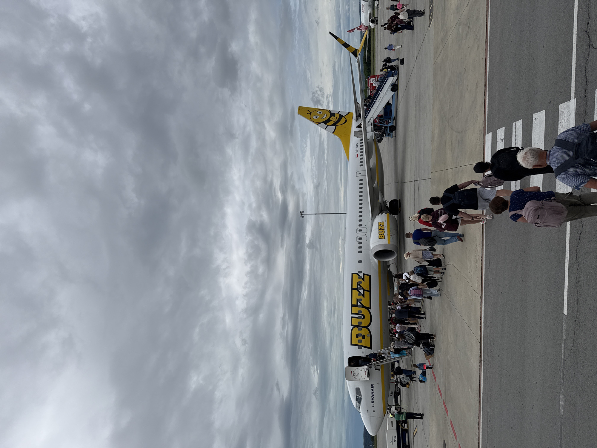 The image depicts an airport tarmac in Aksakovo, Bulgaria, under a cloudy sky. A Buzz airline airplane, branded with a prominent yellow "BUZZ" logo and a bee graphic on its tail, is the central subject. Passengers are seen boarding the aircraft via a mobile staircase. Several people are walking on the tarmac, some with luggage. In the background, another airplane, possibly an Electra, is visible. The scene captures the activity of passengers disembarking or boarding a flight.