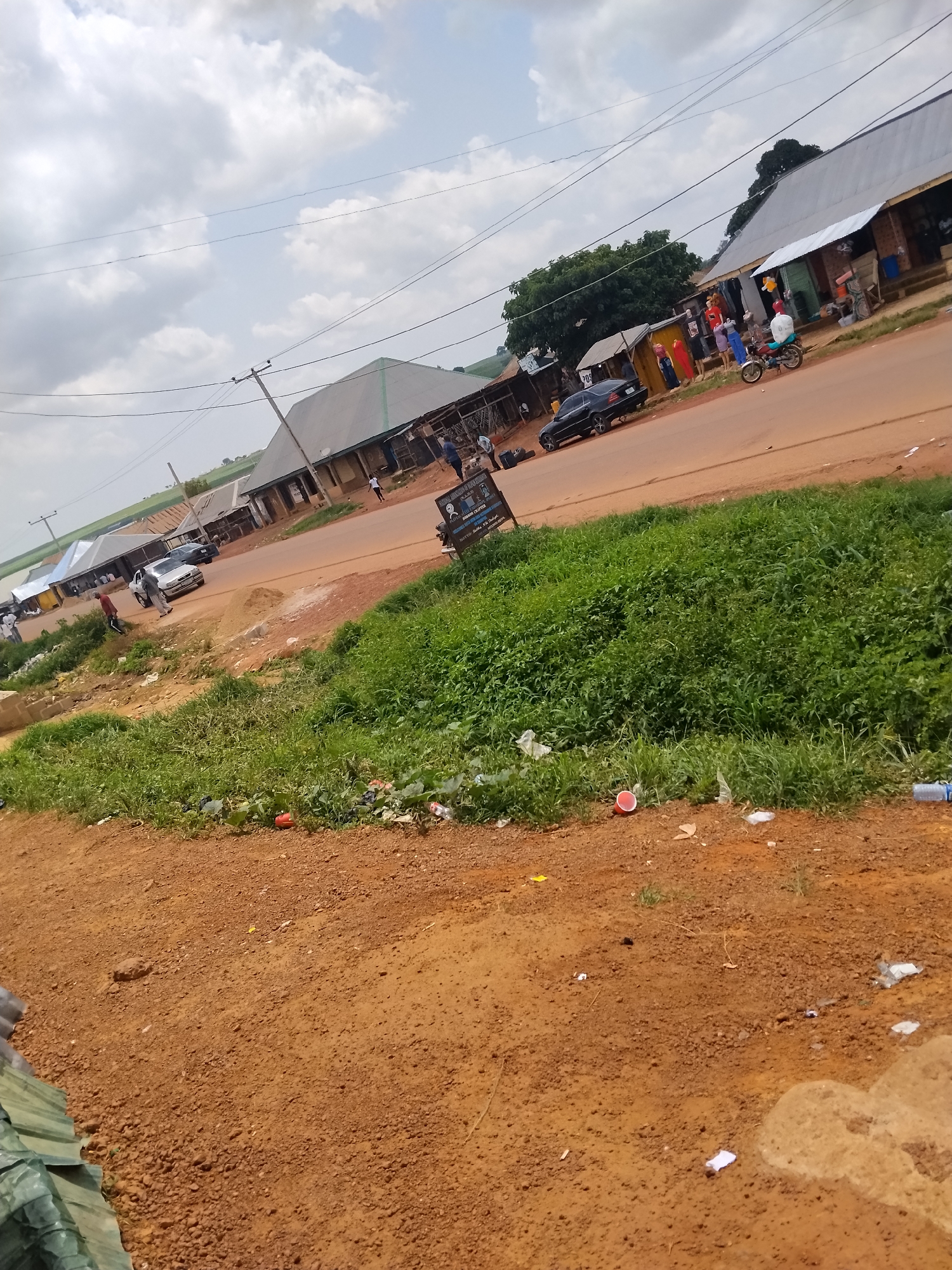 The image depicts a street scene in Durumi, Nigeria, characterized by a wide, unpaved red dirt road lined with shops and residential structures. The sky is cloudy. On the right side of the road, several single-story buildings with corrugated metal roofs house various businesses, including what appears to be a clothing store with mannequins displaying garments. A person is seen riding a motorcycle in front of these establishments. Further down the road, a black car is parked near another shop. On the left side of the road, more buildings and some vegetation are visible. The foreground consists of a dirt embankment covered in patchy green grass and scattered debris, including plastic bottles and red cups, indicating a less-than-pristine environment. Several individuals are present, some walking along the road and in front of the shops. The overall impression is that of a bustling, yet somewhat unkempt, commercial and residential area.