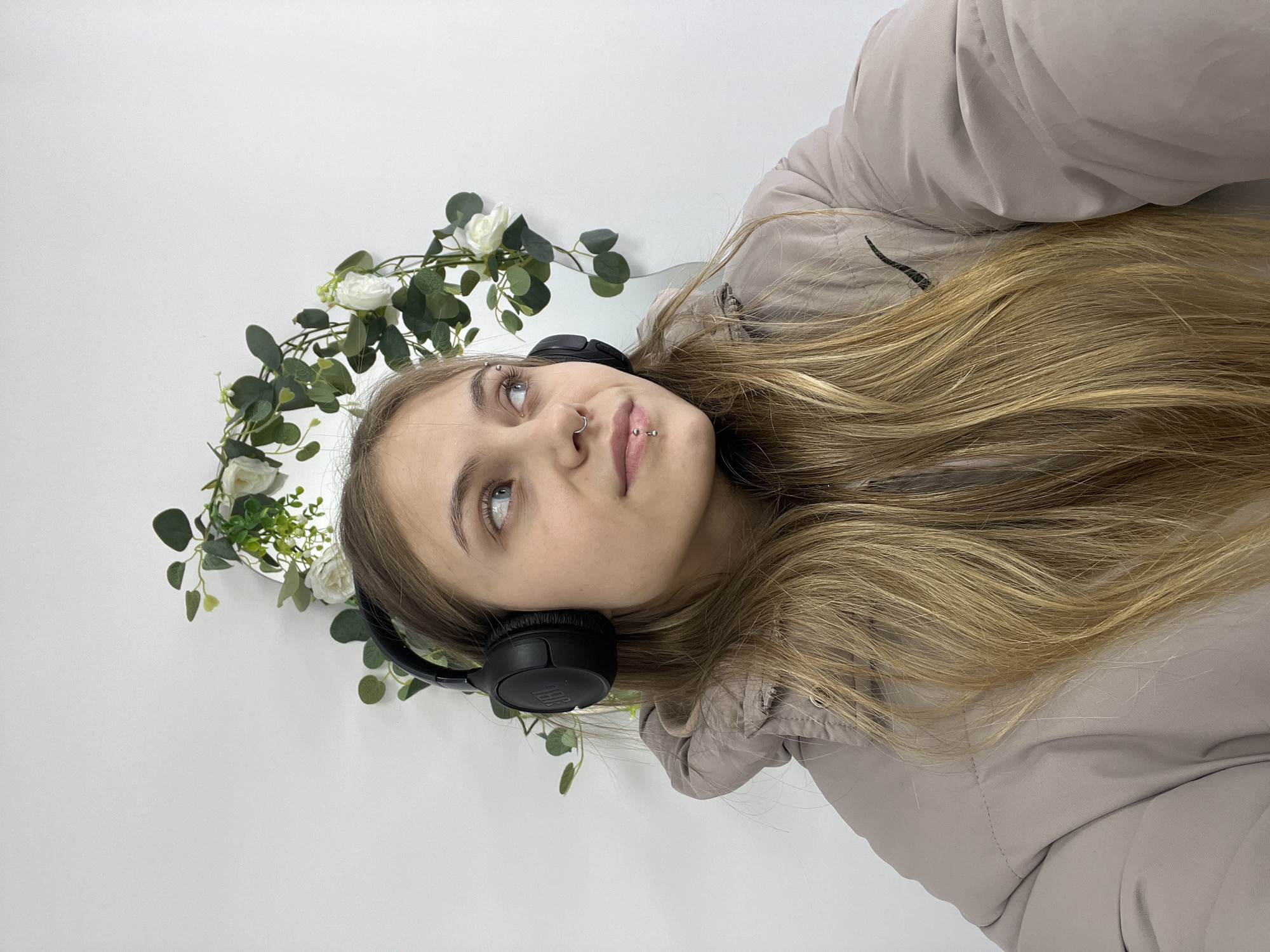 A young woman with light blonde hair is wearing black headphones and a beige puffer jacket, gazing upward and to the left. Her face features a nose ring and a lip piercing. She is positioned against a white wall, adorned with a floral wreath. The image gives no indication of being in or near the city of Фоки, Russia.