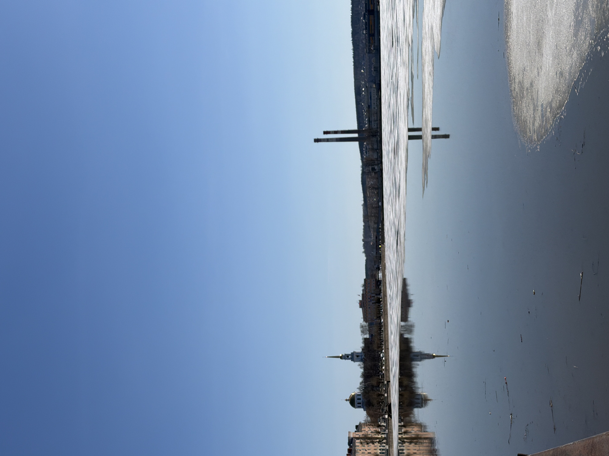 This is a landscape photograph depicting a wide, tranquil body of water, likely a river or reservoir, under a vast, clear blue sky. The image is oriented vertically, with the sky on the left and the water on the right, but it appears to be a horizontally taken shot rotated 90 degrees counter-clockwise.
When viewed in its intended horizontal orientation, the top portion of the frame is dominated by an unbroken, vibrant blue sky. The lower half features the water, which is partially covered with large, melting sheets of ice, indicating a season of thaw, likely late winter or early spring. The water's surface acts as a mirror, producing clear reflections of the distant cityscape and sky.
On the far bank, a town or city stretches along the horizon. Notable structures include a prominent church with a golden dome and a slender spire, positioned towards the left side of the bank. Further along the bank, to the right, two tall, slender industrial chimneys stand out against the sky. Various other buildings and sparse trees line the bank. All these elements are sharply reflected in the calm waters below, creating a symmetrical composition.
There are no people visible in the image. The time of day appears to be daytime, under bright, clear sunlight. The weather is sunny and crisp, suggested by the brilliant blue sky and the presence of melting ice. The scene portrays a serene and somewhat stark landscape, characteristic of the changing seasons in Votkinsk, Russia. No discernible text is visible within the frame.
