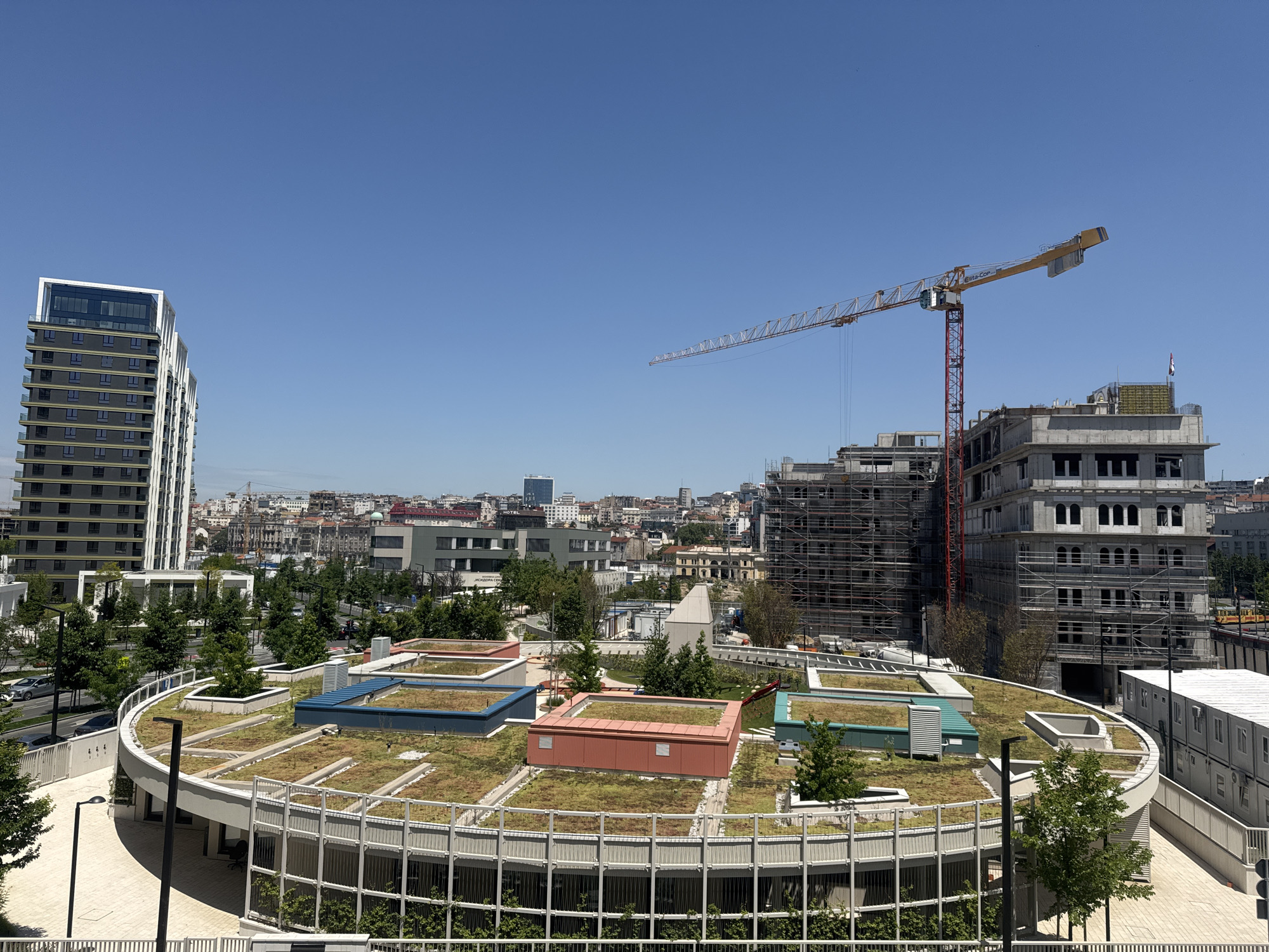 The image depicts a construction site in Belgrade, Serbia, with a clear blue sky overhead. In the foreground, a circular building with a green roof is visible. The green roof features several rectangular structures of various colors, including red and blue, and is dotted with trees. Surrounding this building is a white metal fence.
In the midground, a tall, modern residential building with a dark facade and white accents stands on the left. To its right, a large construction site is in progress, dominated by a red and yellow construction crane. Several multi-story buildings are under construction, covered in scaffolding. In the background, the cityscape of Belgrade unfolds with a mix of older and newer architecture.
The environment appears to be a sunny day, as indicated by the bright lighting and lack of shadows suggesting a high sun position. There are no people visible in the image. The scene is characterized by ongoing urban development and modern architectural elements.