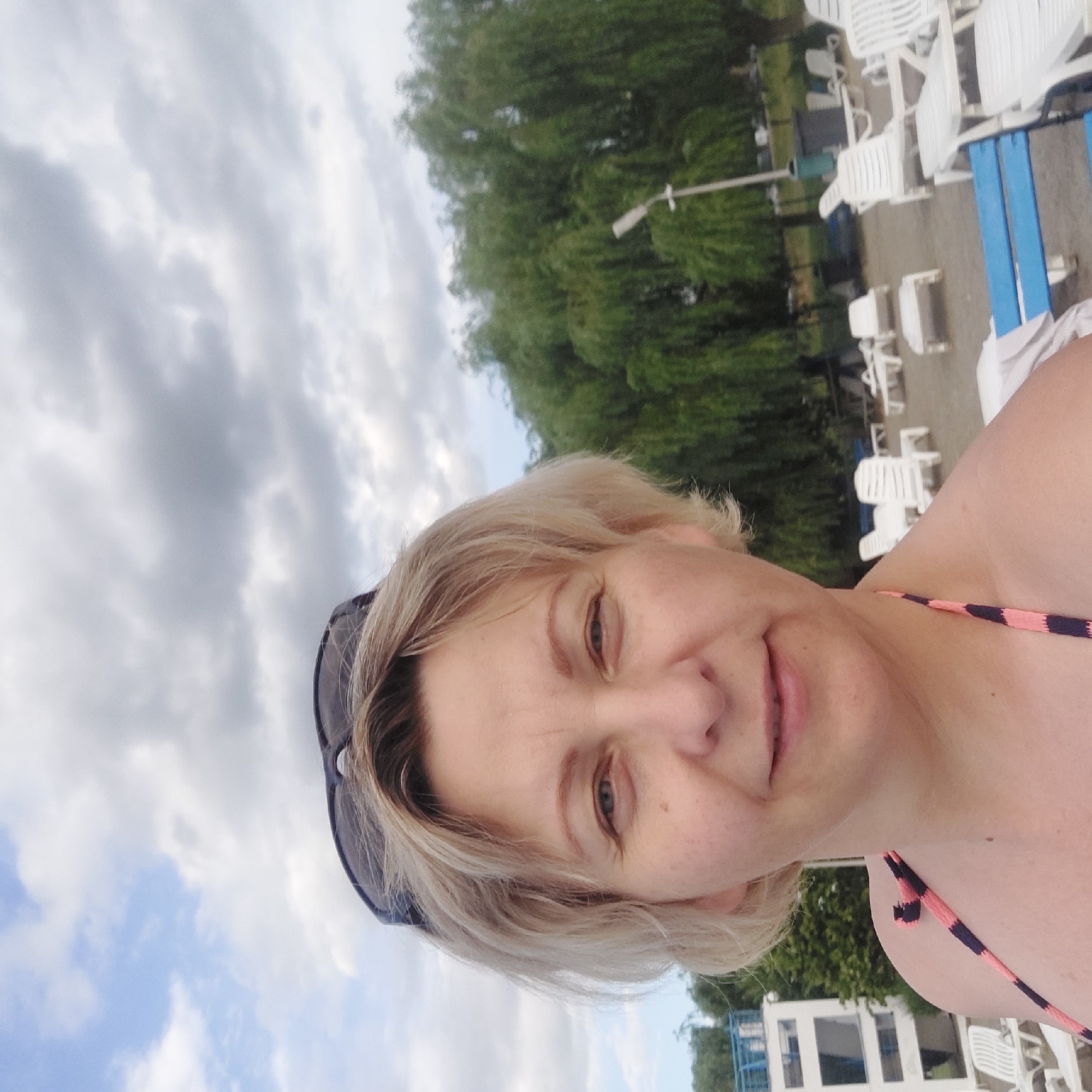 A woman with blonde hair and sunglasses is taking a selfie outdoors. She is wearing a bikini with pink and dark blue stripes. In the background, there are trees, a lamppost, lounge chairs, and a building. The scene appears to be at a beach or a poolside area. The location is not specifically identifiable as Iași, Romania.