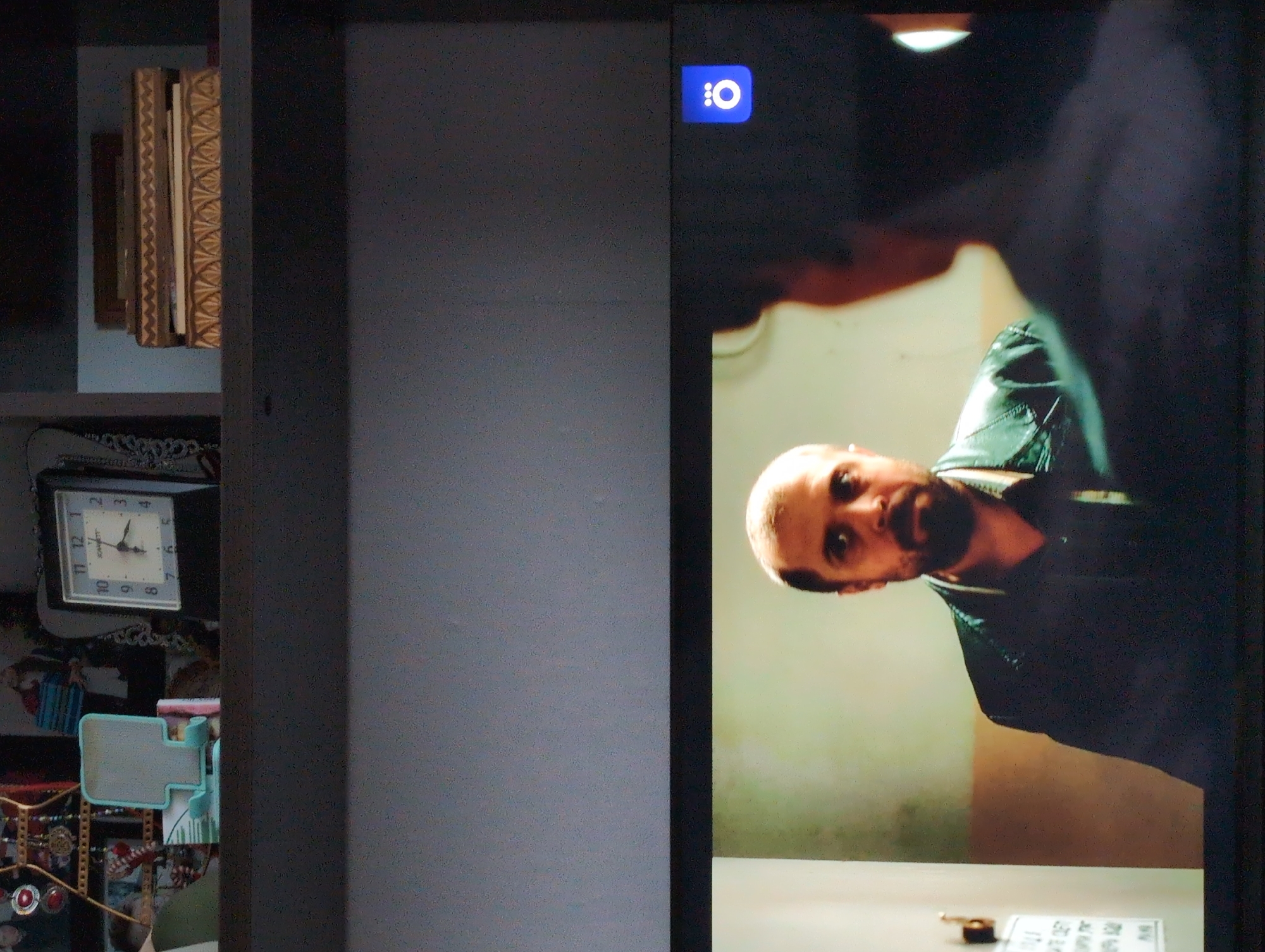 The image shows a television screen displaying a man with a beard and bald head, wearing a black jacket. The background behind him is blurred and indistinct, but appears to be a room with light-colored walls. The television screen also shows the logo of a Russian streaming service. To the left of the television is a shelf containing books, a clock, and various decorative items. There is no indication of the location in the city of Izhevsk, Russia.