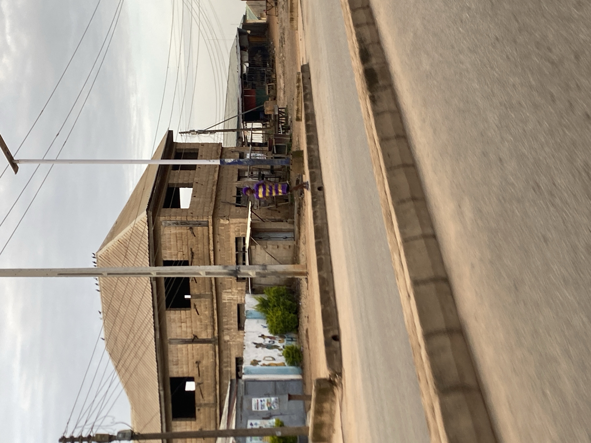 The image depicts an unfinished building in Barakin Lerawi, Nigeria. The structure, made of cinder blocks, has an exposed roof and open window frames. Electrical wires crisscross the scene, strung between utility poles and the building itself. In front of the building, a mural is visible, featuring abstract figures and some greenery. A paved road runs along the right side of the image, with a concrete curb separating it from the building. The sky is overcast, suggesting a cloudy or possibly rainy day. The overall impression is one of ongoing construction and daily life in a Nigerian town. No specific activity or event is clearly discernible, but the presence of the building and the road implies a populated area. The image is oriented vertically, with the road running diagonally from the bottom right to the top left.