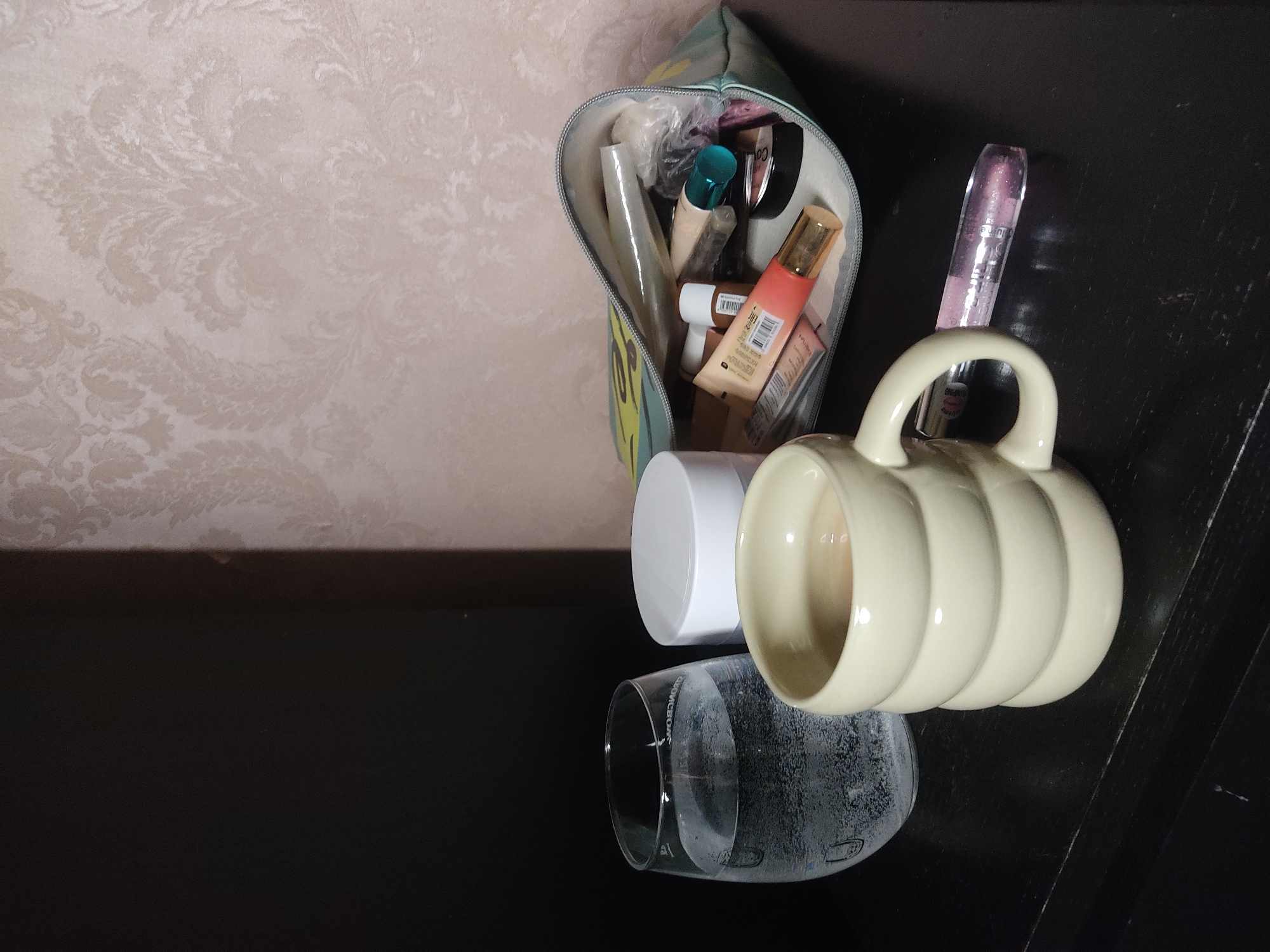The image shows a close-up shot of a dark surface, possibly a table, with several objects on it. There is a makeup bag containing various cosmetic items such as tubes and containers. Next to the bag, there's a clear glass with water condensation on its surface, a white container, and a light-colored mug with a handle. The background includes a patterned wall. The location is unspecified.