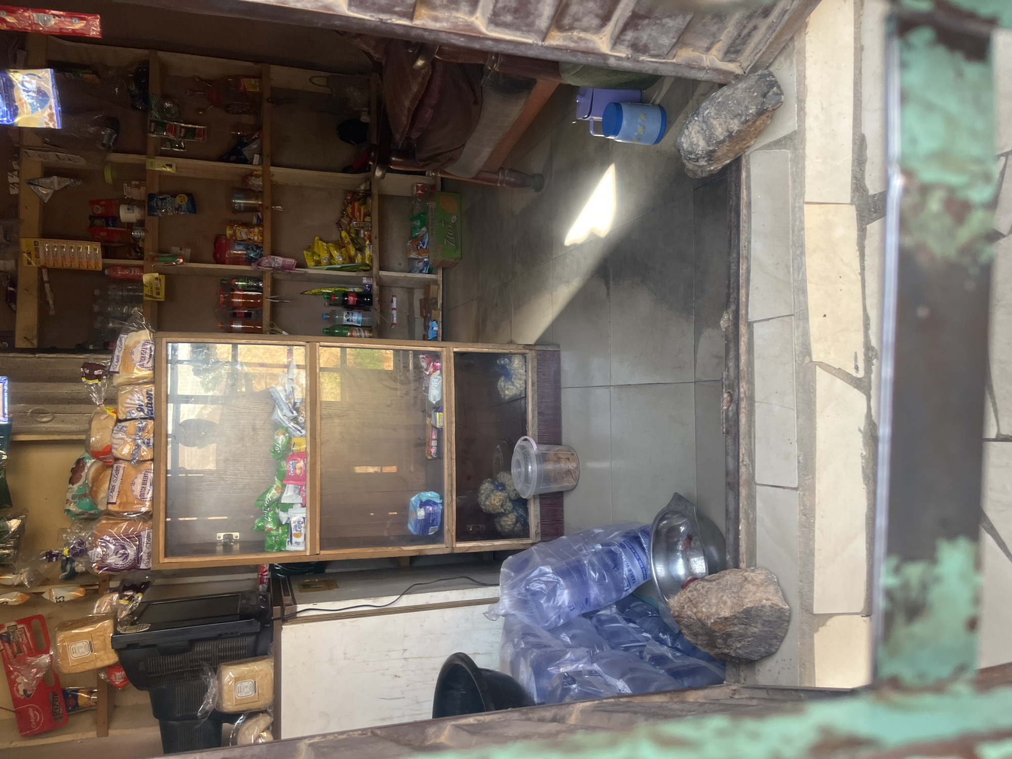 The image depicts the interior of a small convenience store, likely a kiosk or a section of a larger market, situated in the city of Jos, Nigeria. The store is densely stocked with various goods, arranged on shelves and displayed in a glass cabinet. Prominently visible are packaged bread loaves, bottled beverages, snacks in colorful packaging, and what appear to be large sealed containers of water. A television set is also present, situated on a shelf to the left. The flooring is tiled, and sunlight streams in from an unseen source, casting a noticeable shadow across the floor. Two large rocks are placed strategically at the entrance, possibly to weigh down an opening or serve as a barrier. The overall impression is one of a modest, locally operated shop offering a range of everyday consumables.