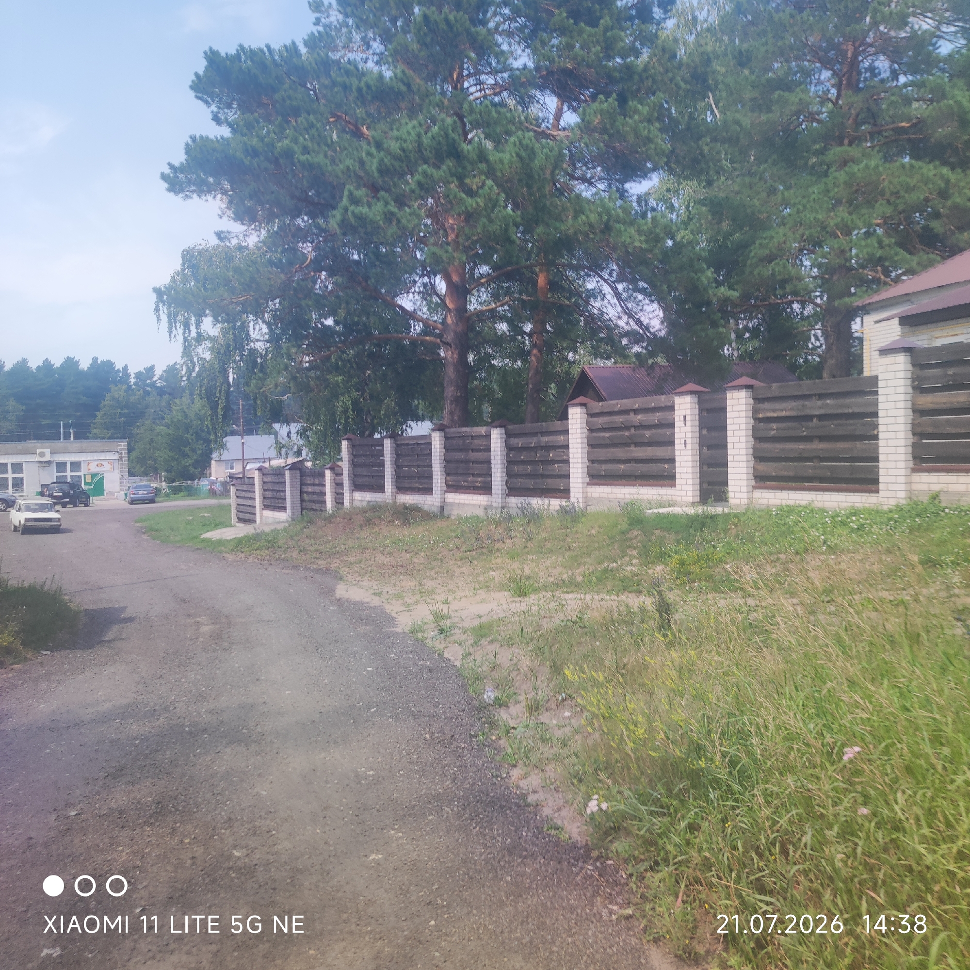 The image shows a dirt road leading towards a building with a white facade and a brown roof. Several cars are parked in front of the building. A wooden fence with brick pillars runs along the side of the road. Behind the fence, there are tall pine trees and other foliage. The sky is partly cloudy. A date and time stamp, "21.07.2026 14:38", and "XIAOMI 11 LITE 5G NE" are visible in the lower part of the image. The scene is located in the city of Баюновские Ключи, Russia.