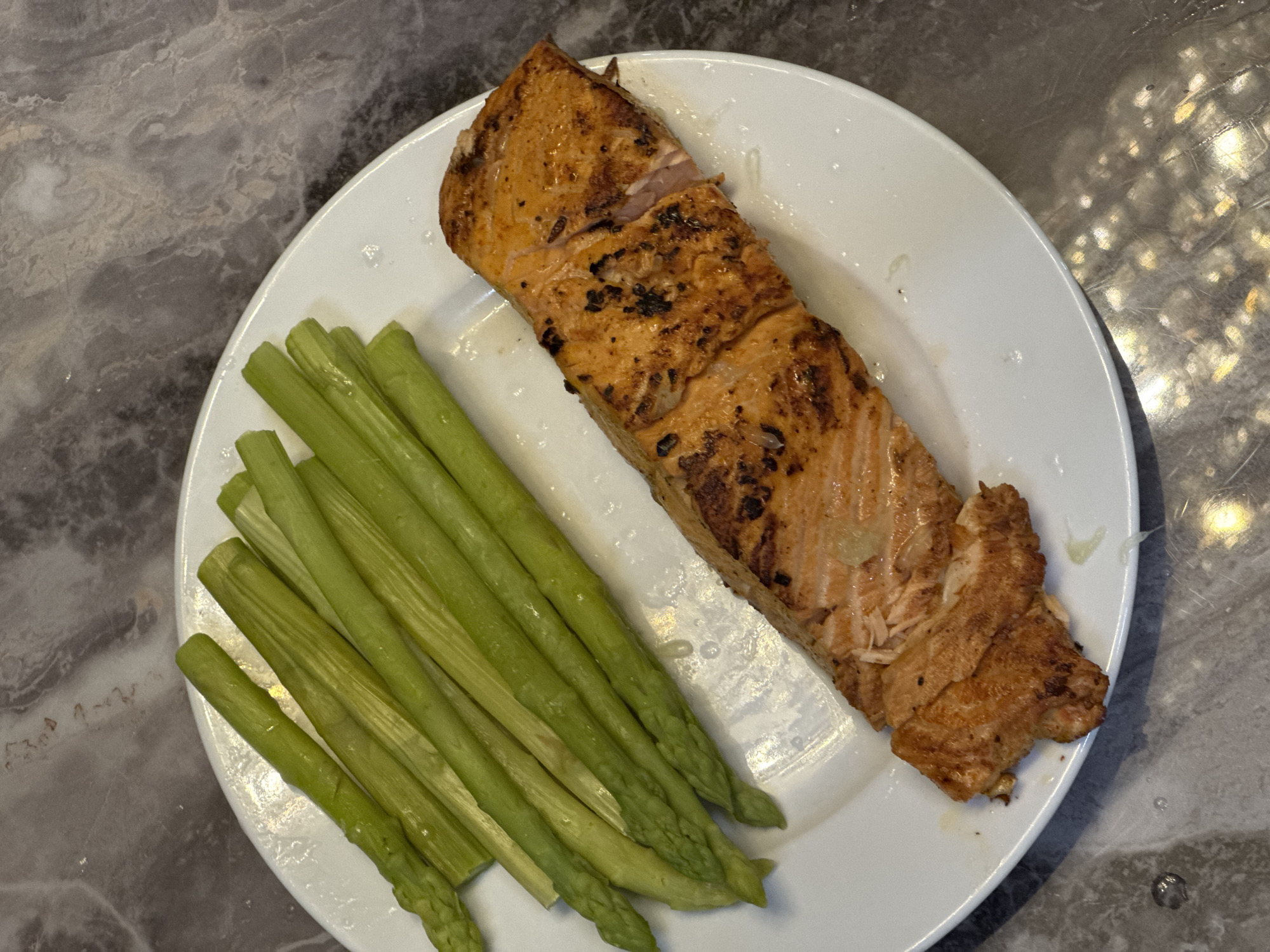 A white plate with grilled salmon and asparagus is displayed on a grey marble countertop. The salmon is golden-brown with grill marks, and the asparagus spears are bright green. The location and city are unknown.
