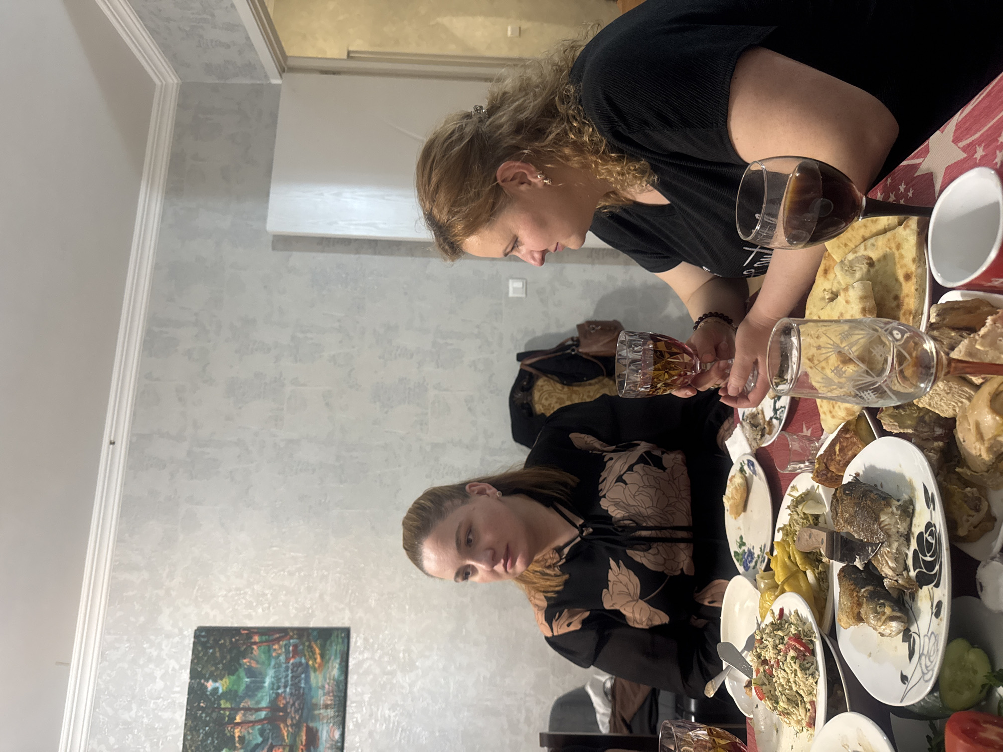 The image captures an indoor scene, likely an evening meal or social gathering, in what appears to be a dining or living room. Two women are seated at a table laden with a variety of food and drinks.
The woman on the right has light brown, curly hair, pulled back with a clip. She is wearing a black ribbed top and is seen looking down towards the table, holding a glass of dark liquid, presumably red wine. She has ear piercings.
The woman on the left has straighter, light brown hair and is wearing a black top with a prominent light-colored floral or leafy pattern. She is also looking down towards the table, engaging with the meal.
The table is covered with a red tablecloth adorned with white star patterns. A diverse spread of food is visible, including plates of fried fish (possibly whole), a salad with what looks like rice or pasta and vegetables, pickled yellow vegetables, and various pieces of flatbread or pastries. Several glasses filled with wine or spirits are on the table, along with an empty white mug. Used plates and cutlery suggest the meal is well underway.
The room's walls are covered in a light-colored, textured wallpaper, possibly light grey or beige. White crown molding is visible along the top of the wall. A framed painting, depicting a landscape with trees and water in an impressionistic style, hangs on the lower left wall. Above the women, a light switch or thermostat is visible on the wall, and an air conditioning unit or shelf is mounted higher up.
The atmosphere appears relaxed and social, characteristic of a family dinner or celebration. Given the context, this gathering likely takes place in Tbilisi, Georgia. There is no visible text in the image.