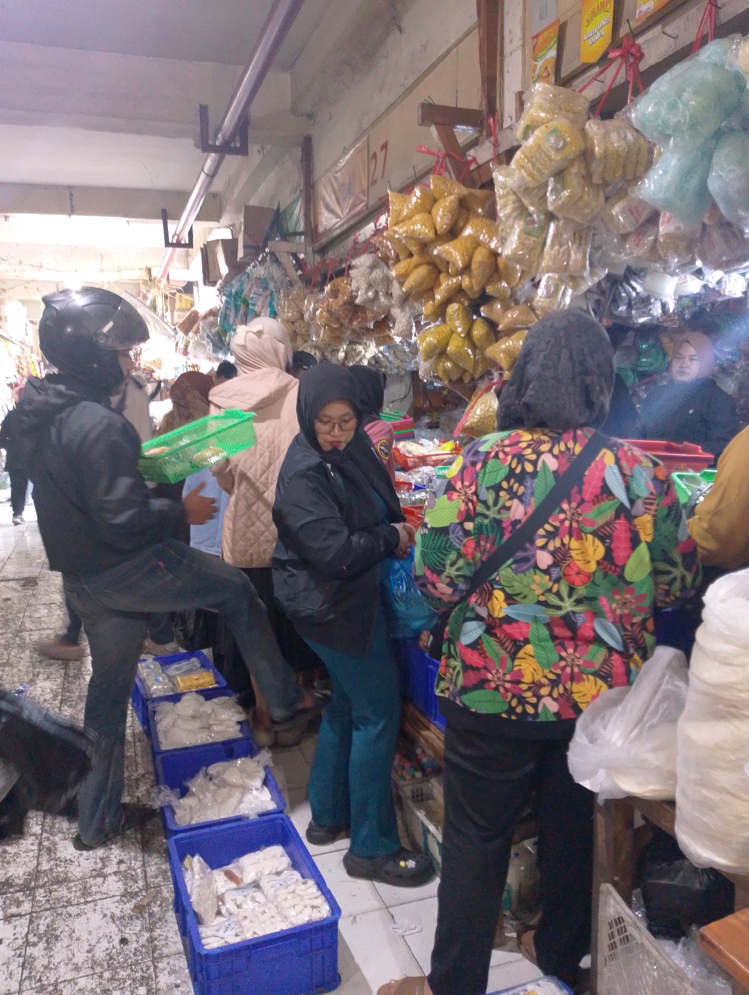 A street market scene is depicted in Lembang, Indonesia, with multiple individuals engaged in shopping or selling. In the foreground, a person wearing a black helmet and jacket is holding a green basket. To their right, a person in a black headscarf and glasses is looking down. Further back, a person in a vibrant floral shirt is facing away from the camera. Various food items, packaged in plastic, are displayed on shelves and hanging from the ceiling, including snacks and dried goods. Blue plastic crates are stacked on the ground, containing more packaged food items. The environment appears to be a covered market with concrete flooring.