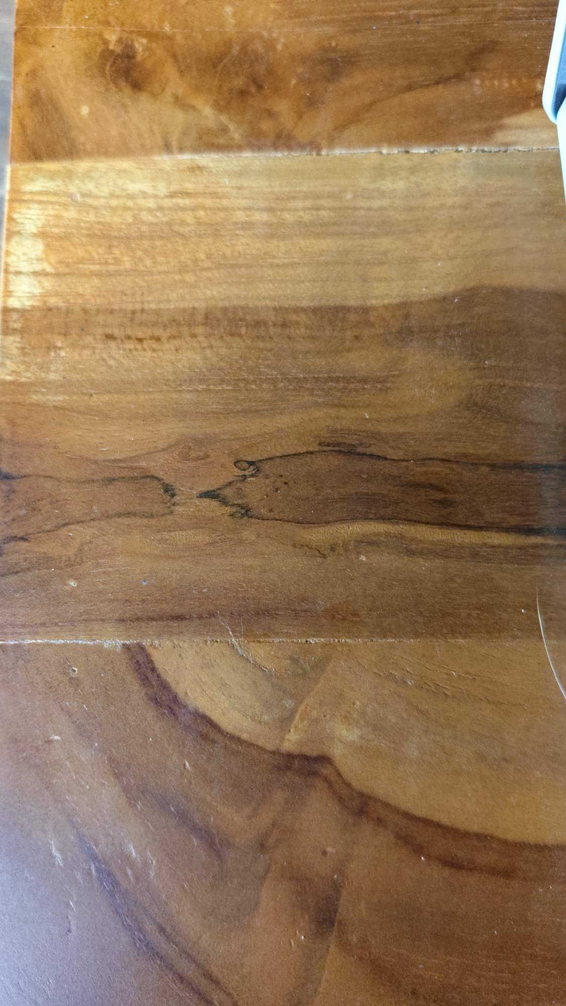 The image displays a close-up, overhead view of a wooden surface, likely a table or countertop. The wood grain is prominent, featuring variations in color from light brown to dark brown, with swirling patterns and darker lines indicating knots or natural imperfections. The surface appears to be polished and slightly reflective. In the upper right corner, a small portion of a blue object, possibly a phone or case, is visible. The lighting suggests an indoor environment, and the overall impression is that of a clean, natural wood material. There are no people or discernible actions taking place. The location is given as Sungai Riam, Indonesia.