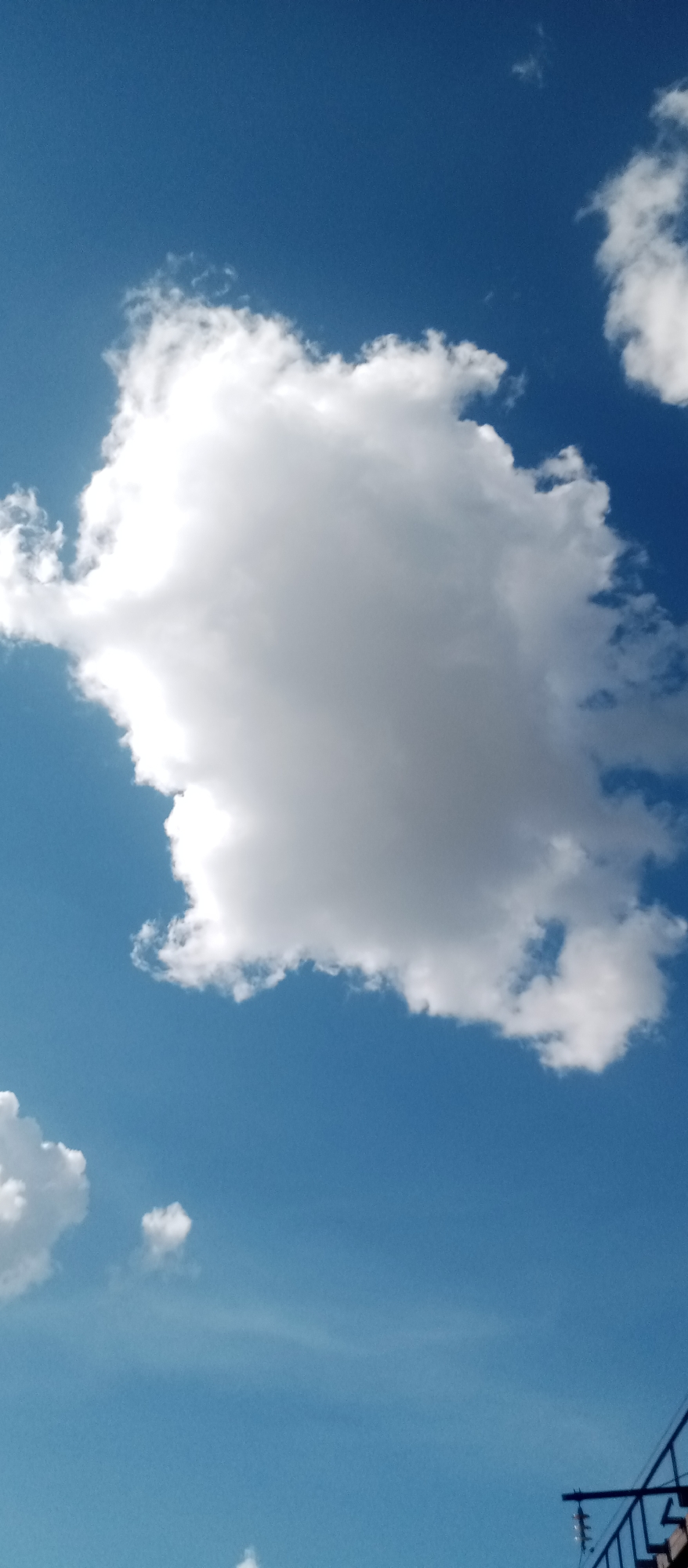 A wide-angle shot captures a bright blue sky filled with fluffy white clouds. The main subject is a large cumulus cloud dominating the center of the frame. Its texture is detailed, with sunlight illuminating its upper portions and casting shadows on its underside, creating a sense of depth and volume. Smaller, scattered clouds dot the sky, some appearing wispy and distant.
In the lower right corner, a sliver of a dark, angular object, likely part of a building or structure with railings and possibly electrical components, intrudes into the frame. This suggests an outdoor setting, possibly a rooftop or elevated vantage point. The clear blue sky and bright illumination of the clouds indicate a sunny day, likely mid-day or afternoon. There is no visible text in the image. The overall mood is serene and expansive, emphasizing the beauty of a clear, cloud-filled sky.