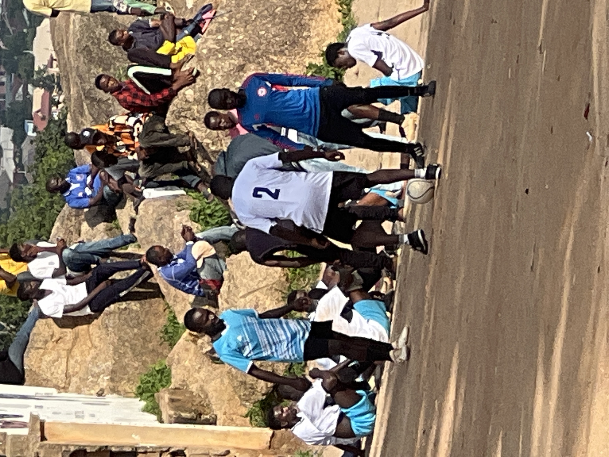 The image depicts an outdoor scene, possibly a training session or informal game of soccer, in Jos, Nigeria. Several young men, mostly in athletic attire, are present. In the foreground, a man in a bright blue and white soccer jersey and black shorts is kicking a soccer ball. Another man in a white jersey with the number "2" on the back is also in the foreground, facing away from the camera. To the left, a man in a blue and white striped jersey and black shorts is visible, looking towards the soccer ball.
In the background, a group of men are seated on large rocks. Some are dressed in casual clothing, while others appear to be spectators. The environment features rocky terrain and some sparse vegetation, suggesting a hilly or elevated area within the city of Jos. The overall impression is one of a casual gathering focused on soccer.