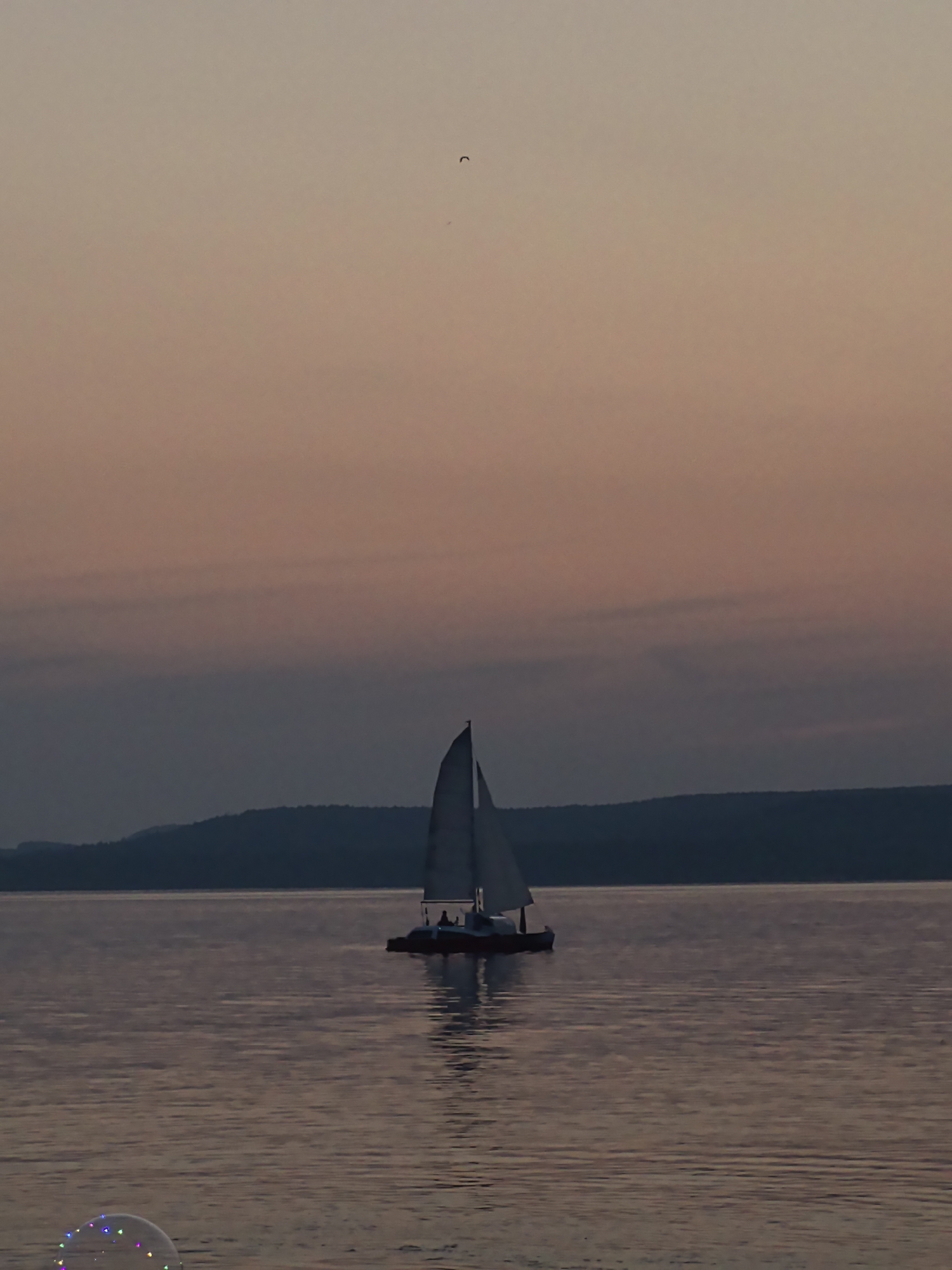 A sailboat with two individuals aboard is visible on calm water at dusk. The sky displays a gradient of soft pink and purple hues. In the foreground, a bubble with colorful lights floats on the water. A distant, dark silhouette of hills borders the horizon.