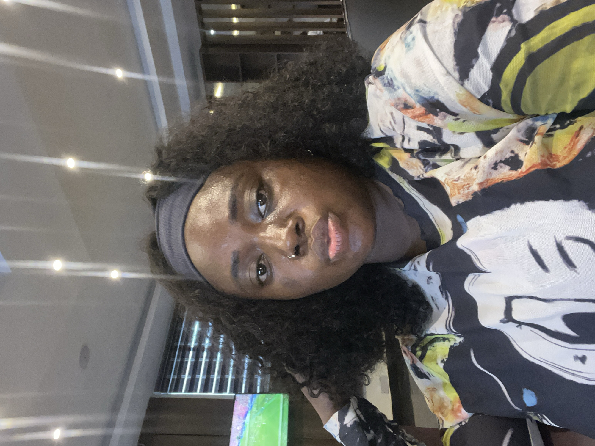 The media file is a close-up, portrait-oriented photo of a young Black woman. She is wearing a grey headband and a colorful, abstract-patterned shirt with black, white, yellow, and orange elements. Her hair is dark, curly, and falls around her shoulders. She has a nose ring and her lips are slightly parted. Her expression is direct and seems calm.
The background suggests an indoor setting with a modern aesthetic. There are bright, recessed lights visible on the ceiling, creating a reflective effect on a light-colored surface, possibly a wall or ceiling. In the mid-ground, a slatted structure, perhaps a divider or part of the building's architecture, is visible. Towards the bottom of the frame, a vibrant green object with multi-colored abstract markings is partially visible, possibly a piece of art or a screen. The lighting in the scene is bright, indicating it is likely daytime or well-lit indoors. The overall mood is casual and personal. There is no visible text. The location context is Abuja, Nigeria.