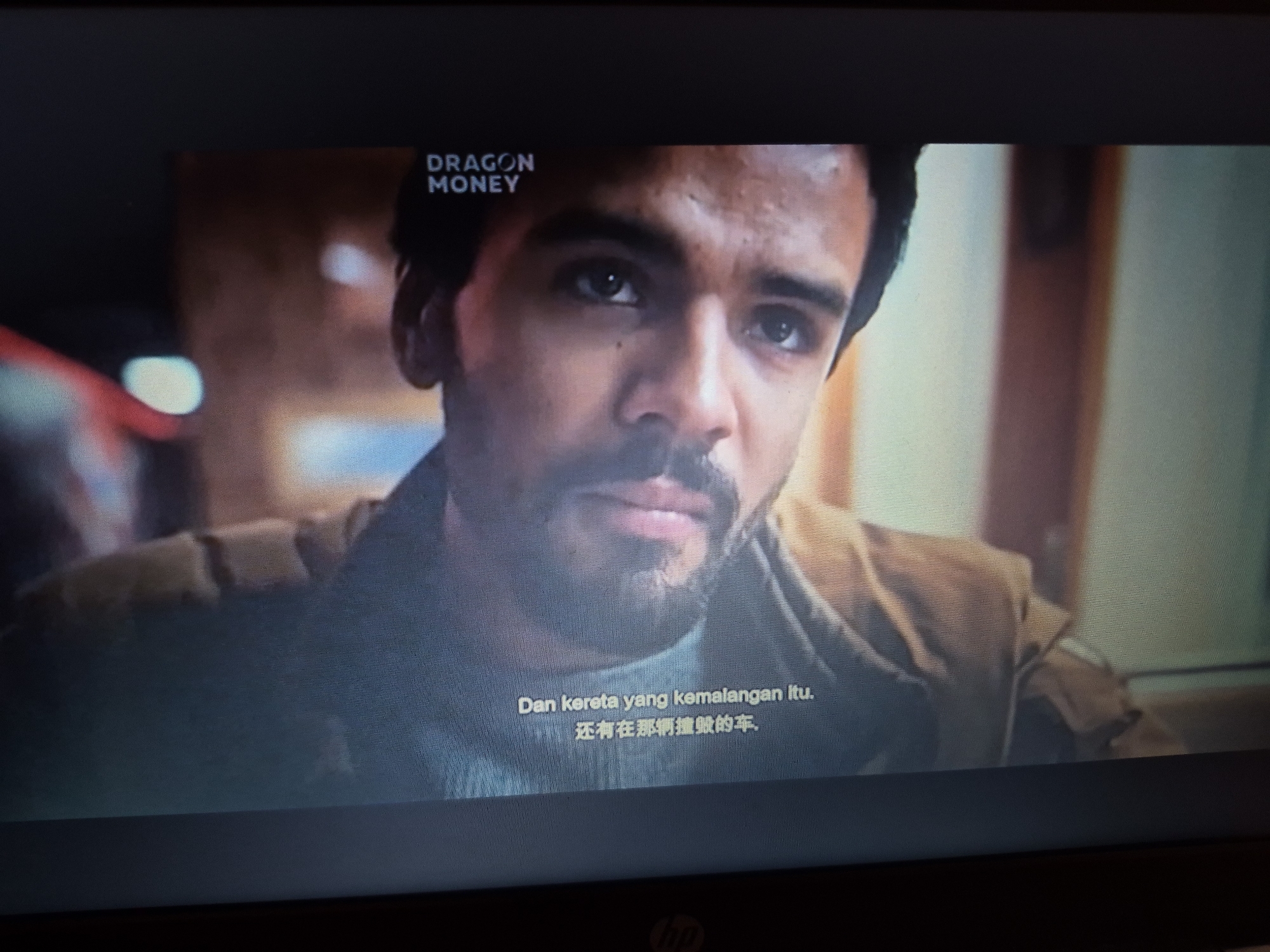 The image shows a close-up of a man's face, looking slightly off-camera. He has dark hair, a mustache, and is wearing a light-colored jacket. Subtitles are visible at the bottom of the screen, displaying text in Malay and Chinese characters. In the top left corner, the words "DRAGON MONEY" are displayed, indicating it is likely a scene from a film or television show. The background is blurred but appears to be an indoor setting with warm lighting. No specific location in Sandwell, United Kingdom, can be determined from the image.