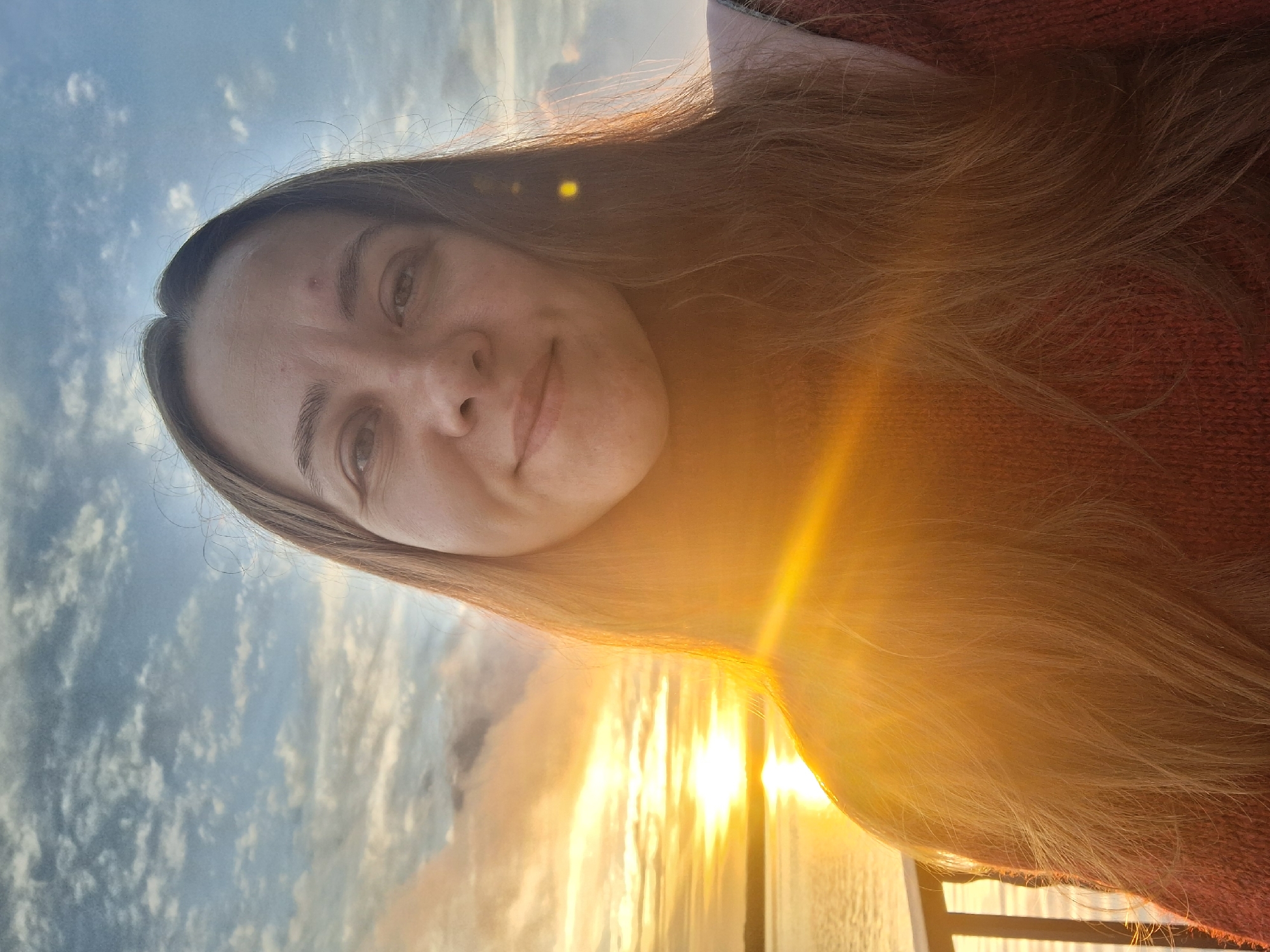 A selfie is taken by a woman with long brown hair, wearing a reddish-orange sweater. She is looking at the camera and smiling. The background shows a body of water and a sunset with clouds in the sky. The sun is low on the horizon and casting a warm glow over the scene. The woman appears to be standing on a deck or balcony with a railing visible in the foreground. The overall impression is one of warmth and tranquility. There is no specific information indicating the location is in Chaikovsky, Russia.