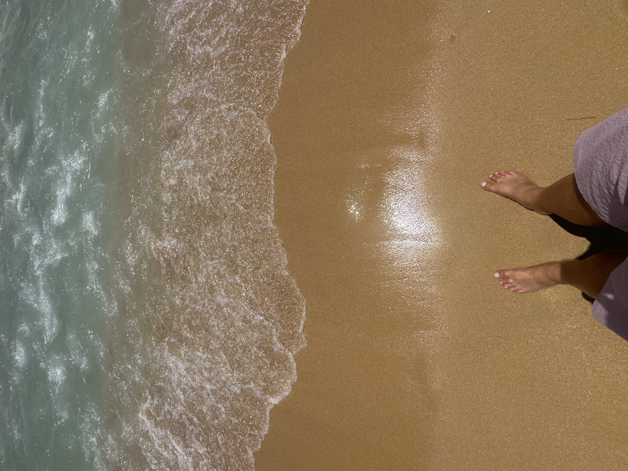 A top-down view of a person's feet standing on a wet, sandy beach. The gentle waves of the ocean are washing up onto the shore, creating white foam. The water is a clear blue-green, and the sand is a warm, golden color, reflecting the sunlight. The person is wearing light-colored shorts, partially visible in the lower right corner. The scene suggests a peaceful and relaxing moment by the sea. The lighting indicates it is daytime, likely sunny given the reflections on the wet sand. The location is identified as Sant Feliu de Guíxols, Spain, a coastal town known for its beaches. There is no visible text in the image.