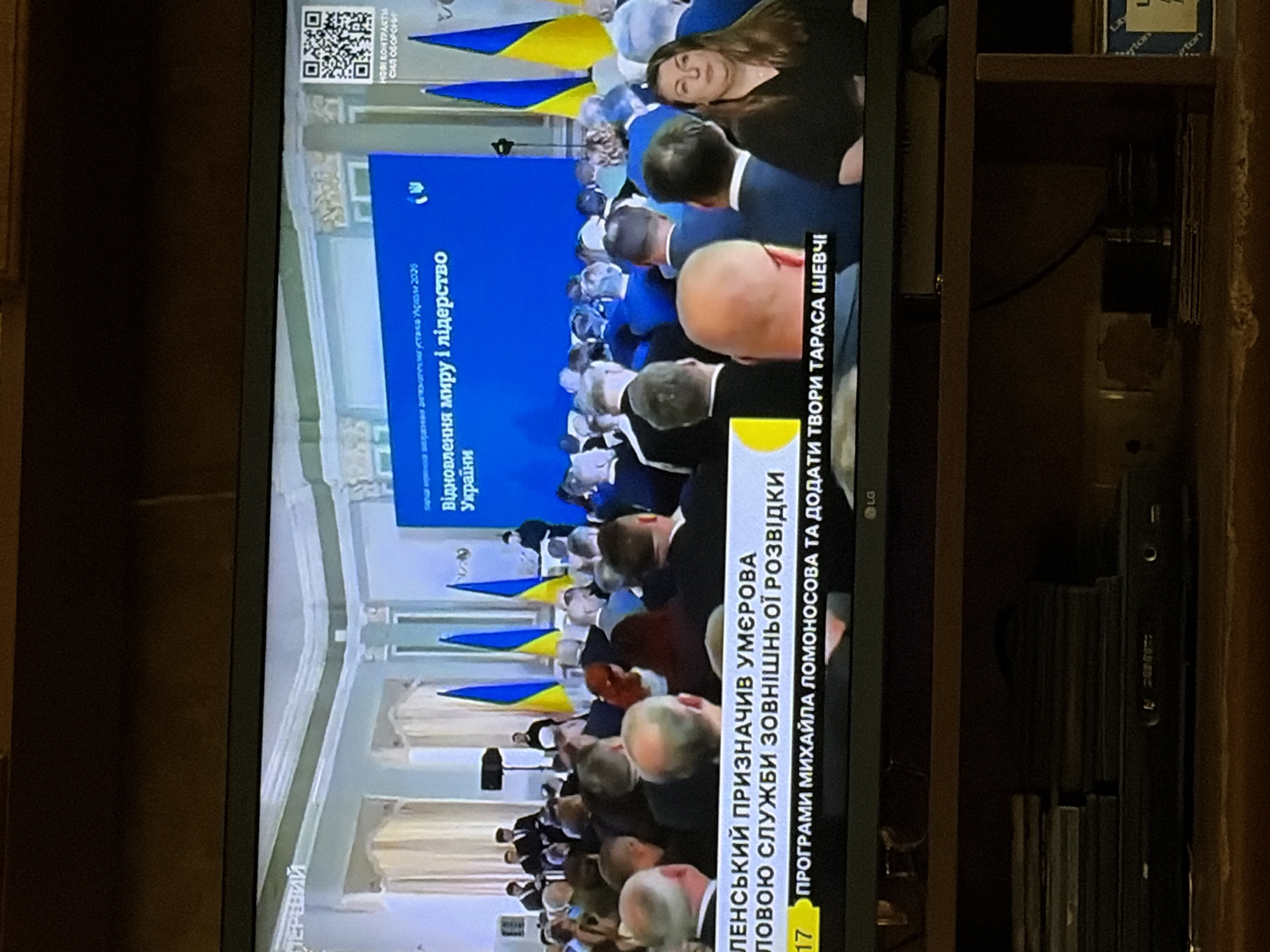 The image shows a television screen displaying a news broadcast. The broadcast features a large blue screen with Ukrainian text, including "ВІДНОВЛЕННЯ МИРУ І ЛІДЕРСТВО УКРАЇНИ" which translates to "Restoration of Peace and Leadership of Ukraine." Below this, there is a ticker tape with Ukrainian text. The scene on the television depicts a formal gathering with many people seated in an audience and several individuals standing on a stage, some holding Ukrainian flags. The image also shows the television itself, which is a flat-screen monitor placed on a wooden shelf. Below the television, there are stacks of DVDs and a black media player. The overall environment suggests an indoor setting, likely a room where the news is being broadcast from.