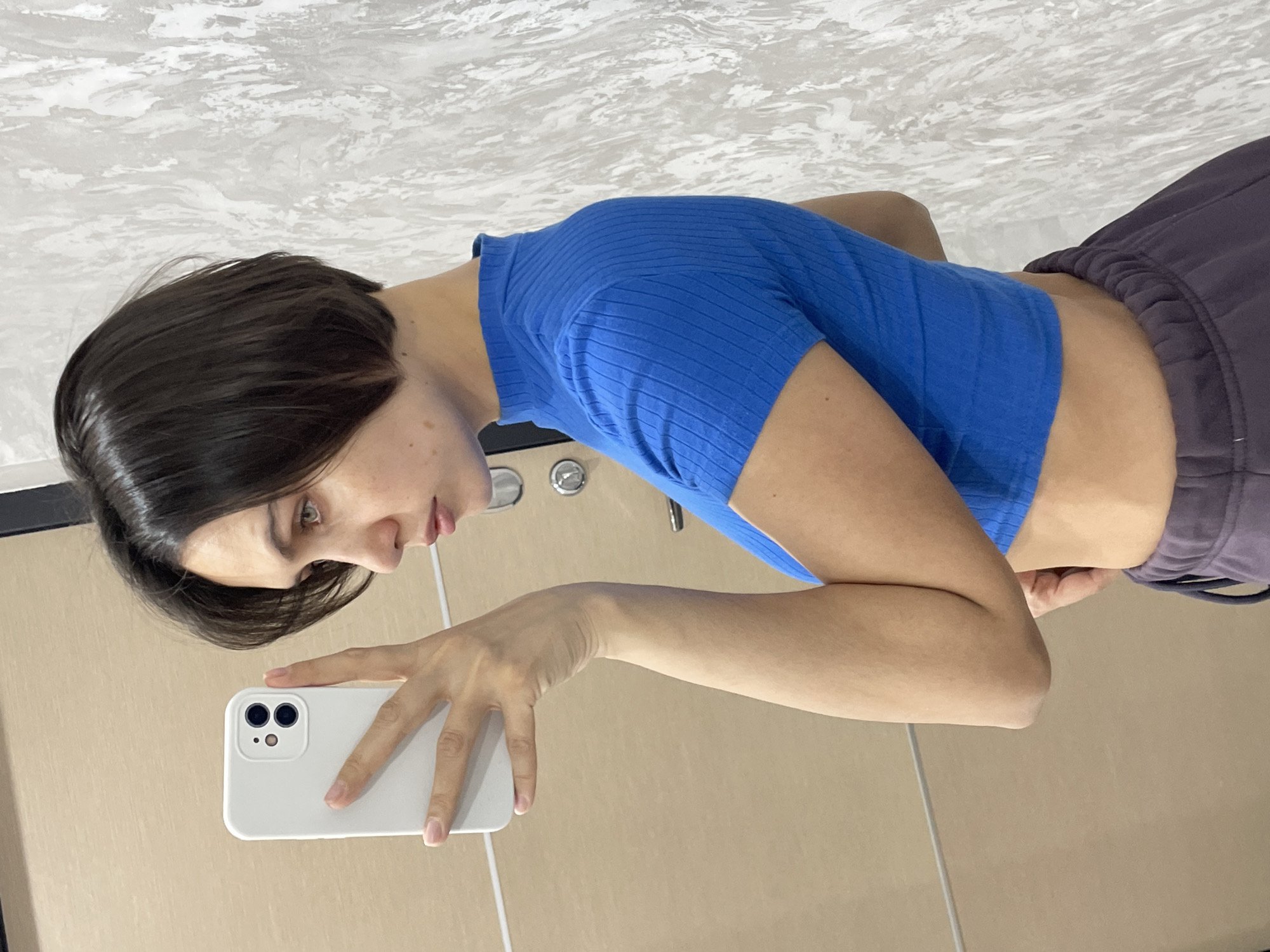 A young woman with short, dark hair is taking a selfie in what appears to be a bathroom. She is wearing a blue ribbed crop top and dark sweatpants. She is holding a white smartphone. The background consists of light-colored tiles and a white countertop with a chrome faucet. No details suggest this photo was taken in Izhevsk, Russia.
