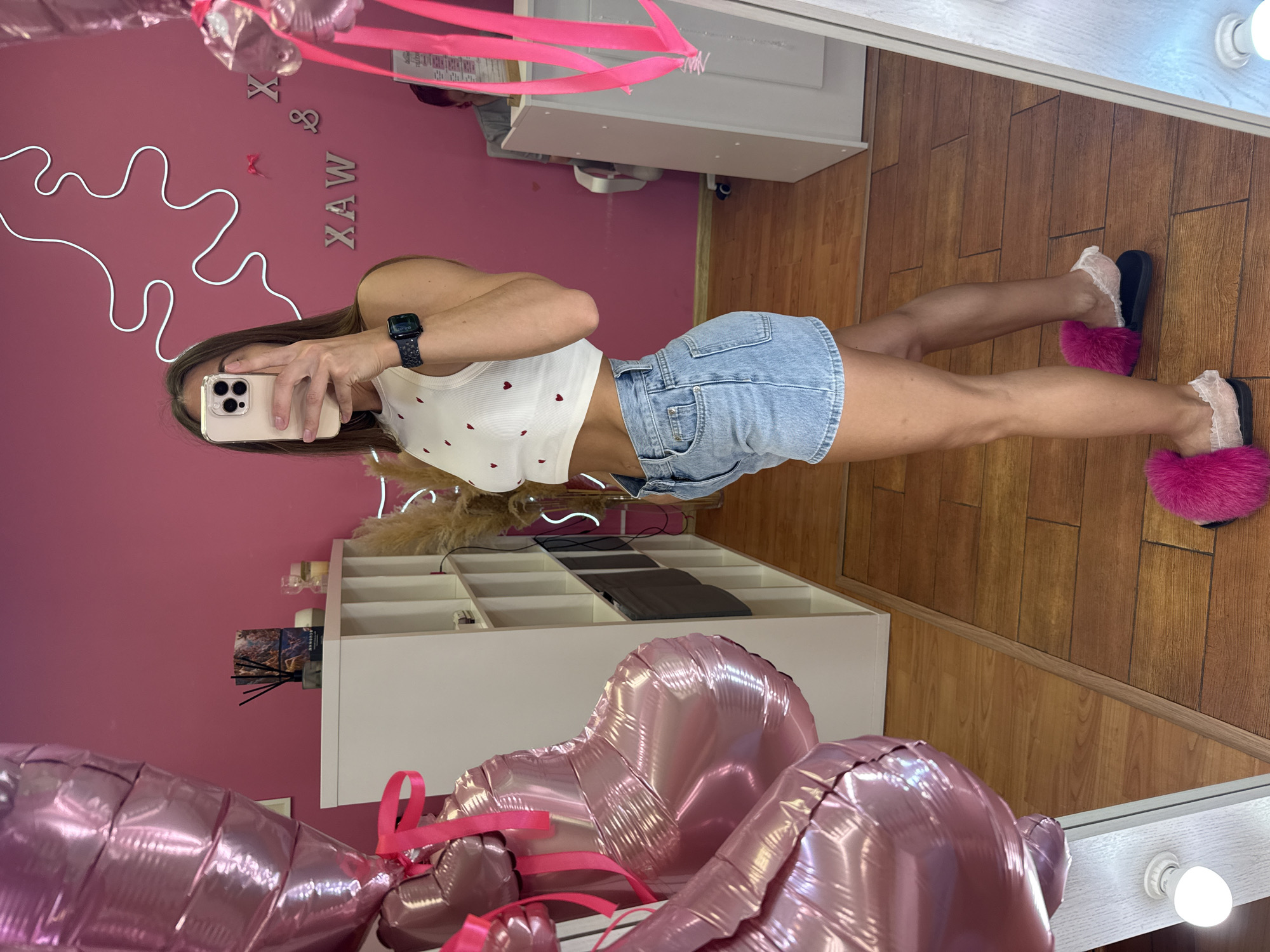 A young woman is taking a mirror selfie in what appears to be a beauty salon in Saint Petersburg, Russia. She is wearing a white crop top with red heart patterns and light-wash denim shorts. She has a smartwatch on her left wrist. Pink heart-shaped balloons are tied to the left of the frame. The walls are painted pink, and there is a white shelf unit to the left of the woman. In the background, there is a person sitting at a desk. The floor is made of wooden planks.