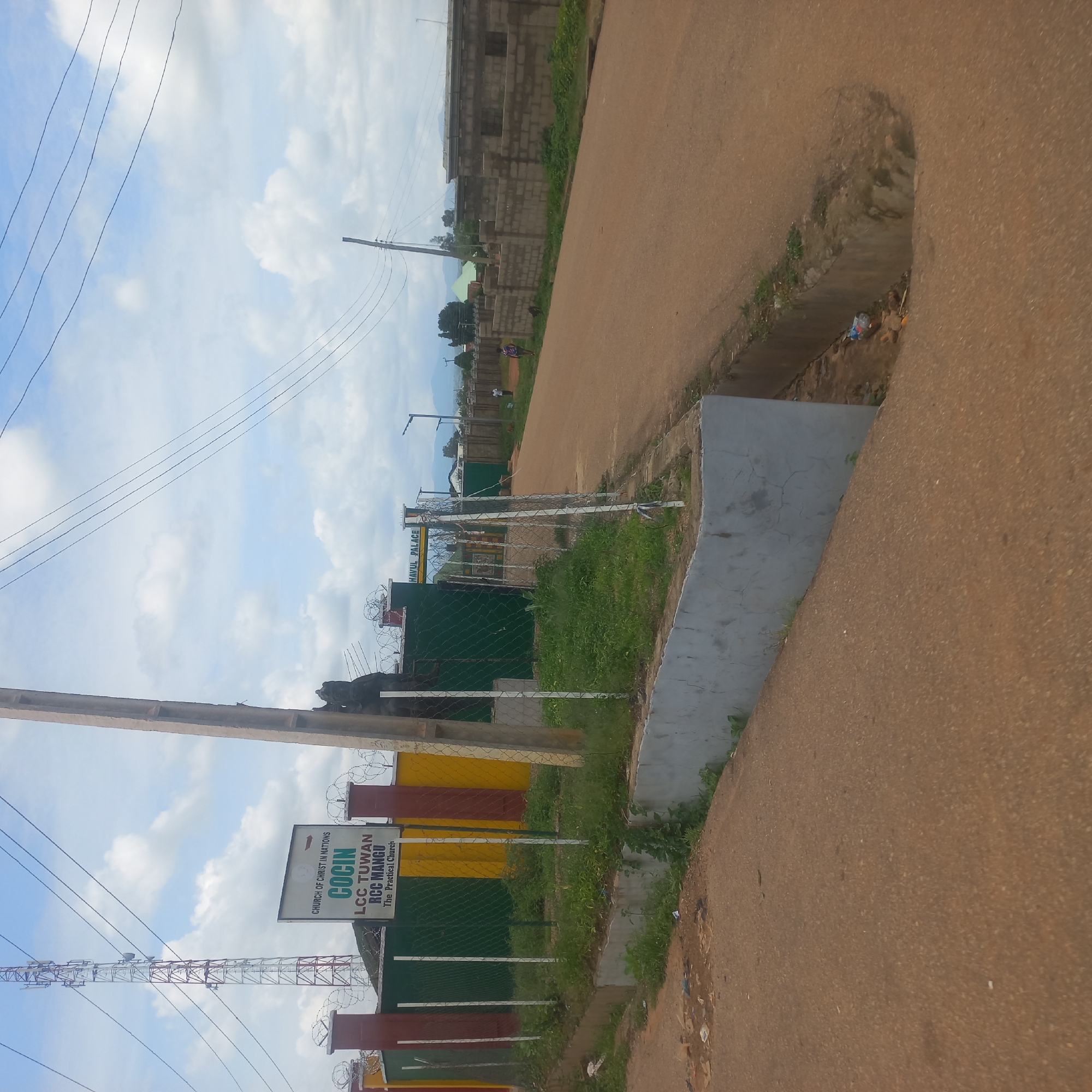 The image depicts a street scene in Mangu Town, Nigeria, under a partly cloudy sky. On the left, a sign identifies the location as "CHURCH OF CHRIST IN NATIONS COÇIN LCC TUWAN RCC MANGU The Practical Church." Adjacent to the sign is a tall telecommunications tower. The church property is enclosed by a fence with red, yellow, and green sections and barbed wire at the top. A concrete drain runs alongside the road on the right, with some debris visible at the bottom. The road itself is unpaved and reddish-brown. In the distance, a wall constructed of concrete blocks lines the right side of the road, and several buildings are visible further back. Two individuals are walking on the road in the middle ground. A utility pole stands prominently in the foreground on the left.