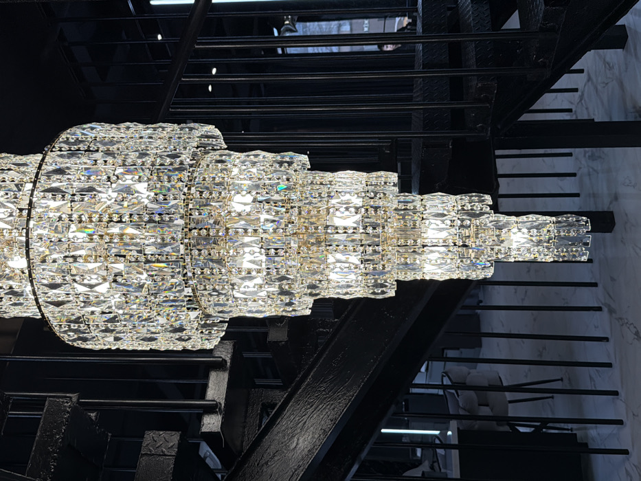 The image displays a modern interior located in Moscow, Russia. A large, multi-tiered crystal chandelier, composed of numerous illuminated, clear, faceted crystal elements, hangs prominently in the center of the frame. To the left and right, a dark, industrial-style metal staircase with black painted diamond plate treads ascends. The floor features a light-colored marble pattern. In the background on the left, contemporary furnishings, including chairs and tables, are visible, alongside vertical LED strip lighting. On the right, a window reveals an exterior environment, with a faint, partial silhouette of an individual discernable through the glass. No significant actions or interactions are occurring.