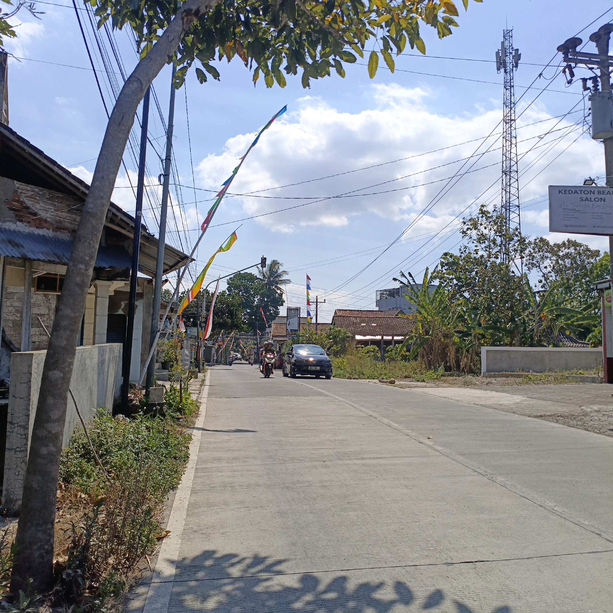 The image depicts a street scene in Salatiga, Indonesia, under a bright blue sky with scattered white clouds. A paved road runs diagonally from the bottom right to the upper left of the frame, with a concrete sidewalk on the left side. A large tree trunk is prominent in the foreground on the left, with its branches and leaves partially obscuring the sky.
Along the sidewalk, there are several buildings with brick walls and corrugated metal roofs, some appearing older and in disrepair. Numerous electrical wires are strung overhead, supported by poles and a prominent telecommunications tower in the mid-ground to the right. Decorative flags in various colors are strung along poles on both sides of the road.
Traffic on the road includes two motorcycles with riders and one dark-colored car. The motorcycles are traveling away from the viewer, while the car is approaching. Vegetation, including banana trees and other plants, is visible on the right side of the road and in the background. A sign for "KEDATON BEAUTY SALON" is partially visible on the right. The scene suggests a typical urban or suburban street in Indonesia on a sunny day.