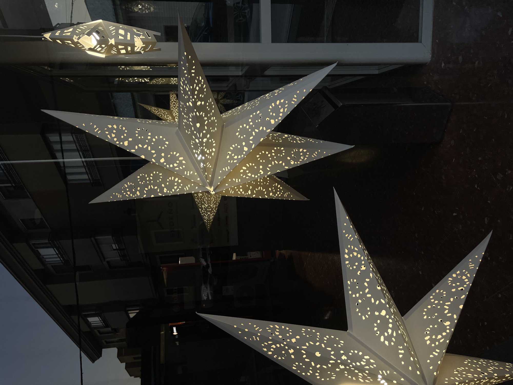 This is a close-up, angled outdoor shot of a storefront window display featuring decorative, illuminated paper stars. The stars are white with intricate cut-out patterns that allow light to shine through, creating a warm glow. In the background, a dark, multi-story building with windows is visible, reflecting the twilight sky. Some reflections of cars and interior lights are also present in the window. The scene is set in La Línea De La Concepción, Spain, a city on the easternmost edge of the Campo de Gibraltar, bordering Gibraltar.