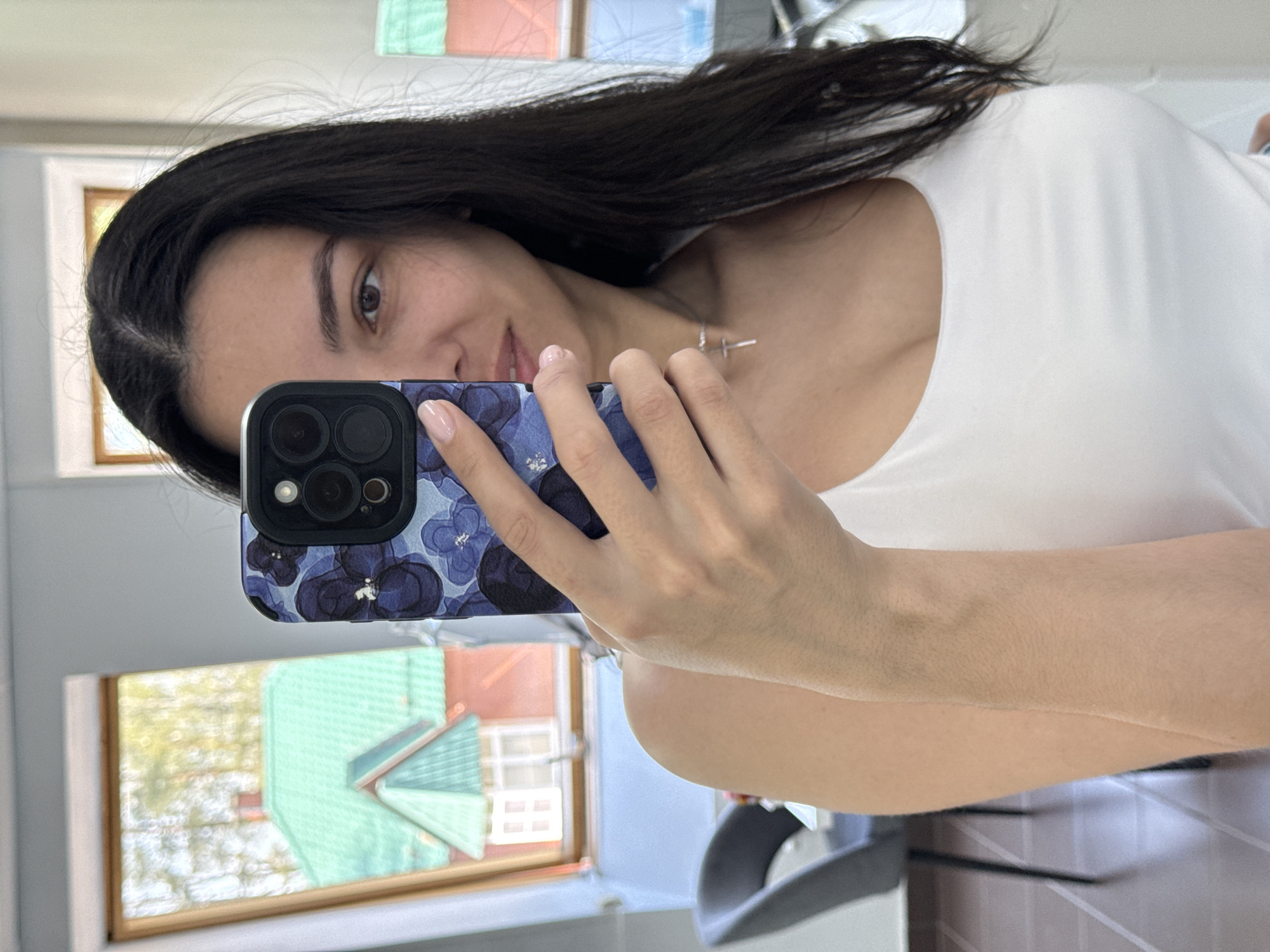 A woman is taking a selfie in what appears to be an indoor setting. She is holding a smartphone with a floral-patterned case. She has dark hair and is wearing a white top. Through a window in the background, a house with a green roof and red siding can be seen. Based on the house architecture, the location is likely Chaikovsky, Russia.