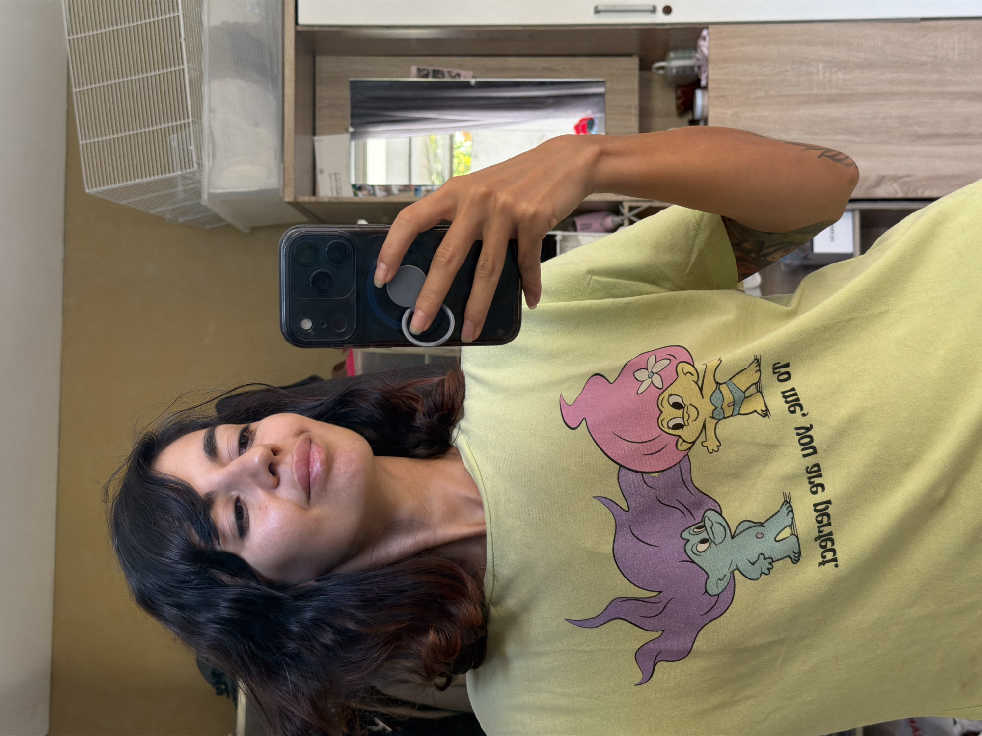 A woman is taking a mirror selfie in Kuta Selatan, Indonesia. She is wearing a light green t-shirt with a cartoon graphic of two trolls. The woman has dark, wavy hair and is looking at her phone, which she is holding in her right hand. In the background, a white hamster cage is visible on top of a shelf. The room appears to be a bedroom or dressing area, with a wardrobe and a window with grey curtains in the reflection.