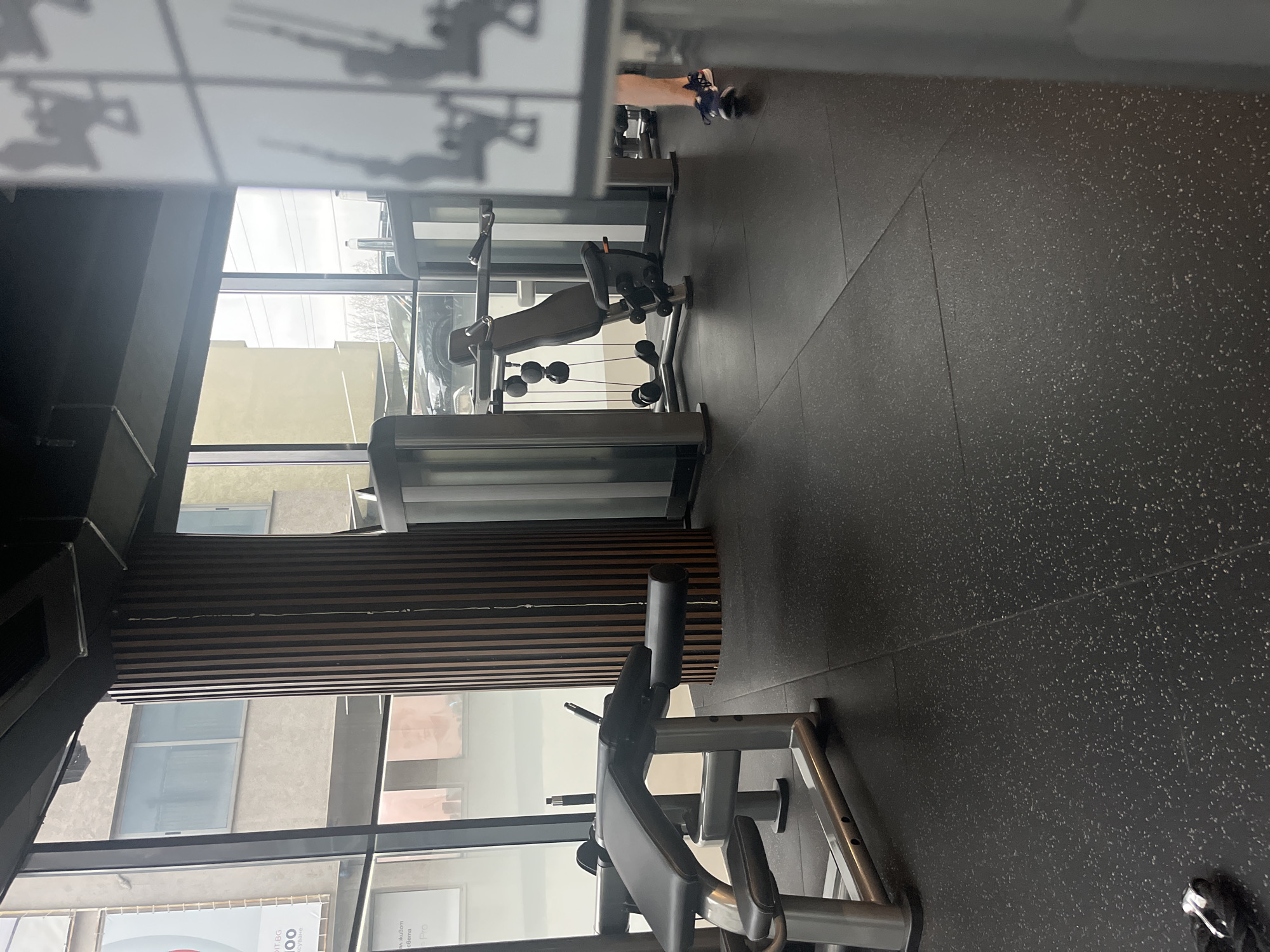 The image depicts a gym interior, potentially located in Sofia, Bulgaria, given the presence of Bulgarian text in the bottom left corner. The gym is modern with large windows offering a view of a building across the street. Gym equipment is visible, including a weight machine and a bench press. The floor is a dark, speckled material, likely rubber or similar gym flooring. The lighting is bright, due to the large windows. There is at least one person in the gym, with their legs and shoes partially visible near the top right of the frame. The gym ceiling has a tiled appearance. The walls have horizontal wooden slats.