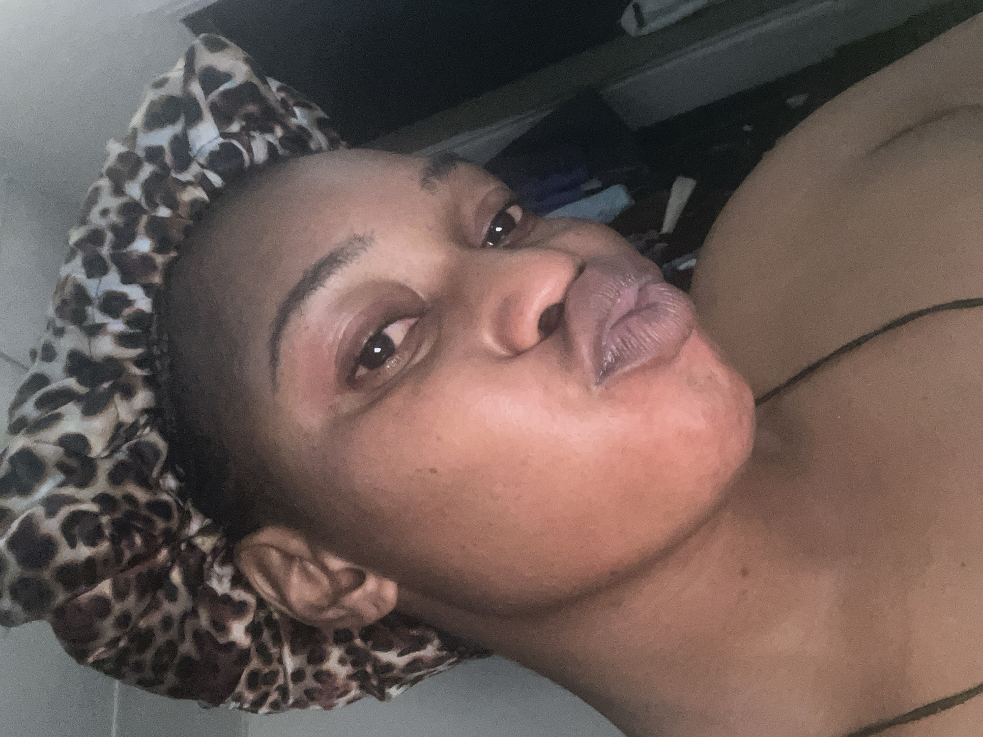 The image shows a close-up, angled selfie of a Black woman wearing a leopard-print bonnet. Her lips are pursed, and she is looking towards the camera. Her skin appears smooth with some subtle redness on her cheek. The background is dark and out of focus, with a hint of what might be clothing or fabric visible on the right side. The lighting is soft, casting a slight glow on her face. This image does not provide any specific visual cues or context to determine its location within Jos, Nigeria, or any other specific city.