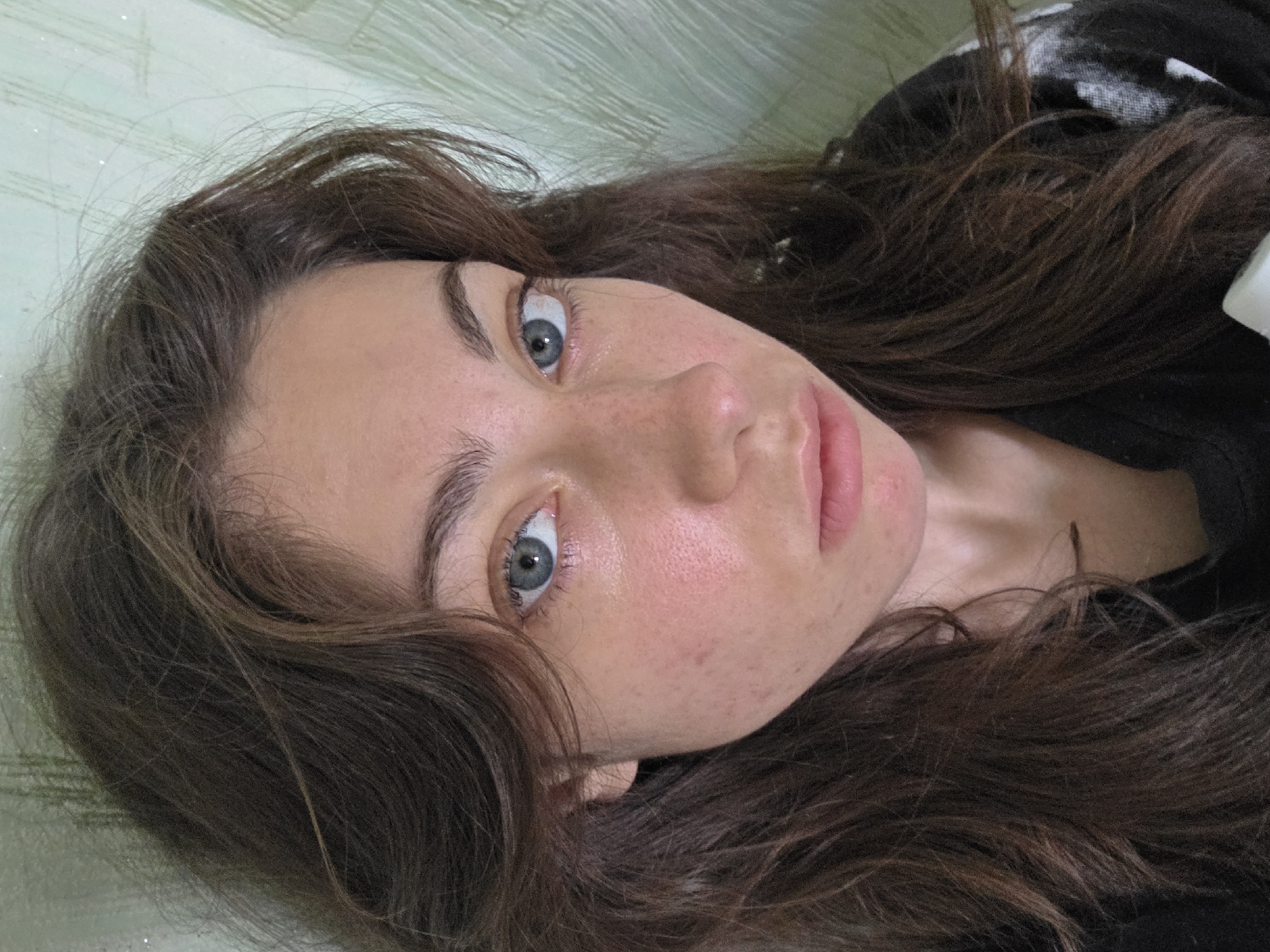 The image is a close-up, head-and-shoulders portrait of a young woman with long, dark brown hair. She has light blue eyes, prominent eyebrows, and fair skin with some visible blemishes and redness on her cheeks. Her lips are closed and appear naturally colored. She is wearing a black top, the pattern of which is partially visible on the right side of her chest. The background is a textured, light green wall, with vertical lines suggesting wallpaper or a similar finish. There are some small, sparkling specks visible in the background, possibly dust or a lighting effect. The overall lighting is soft, with a slight sheen on the woman's face indicating natural light. There is no discernible city location, including Chita, Russia, indicated in the image.