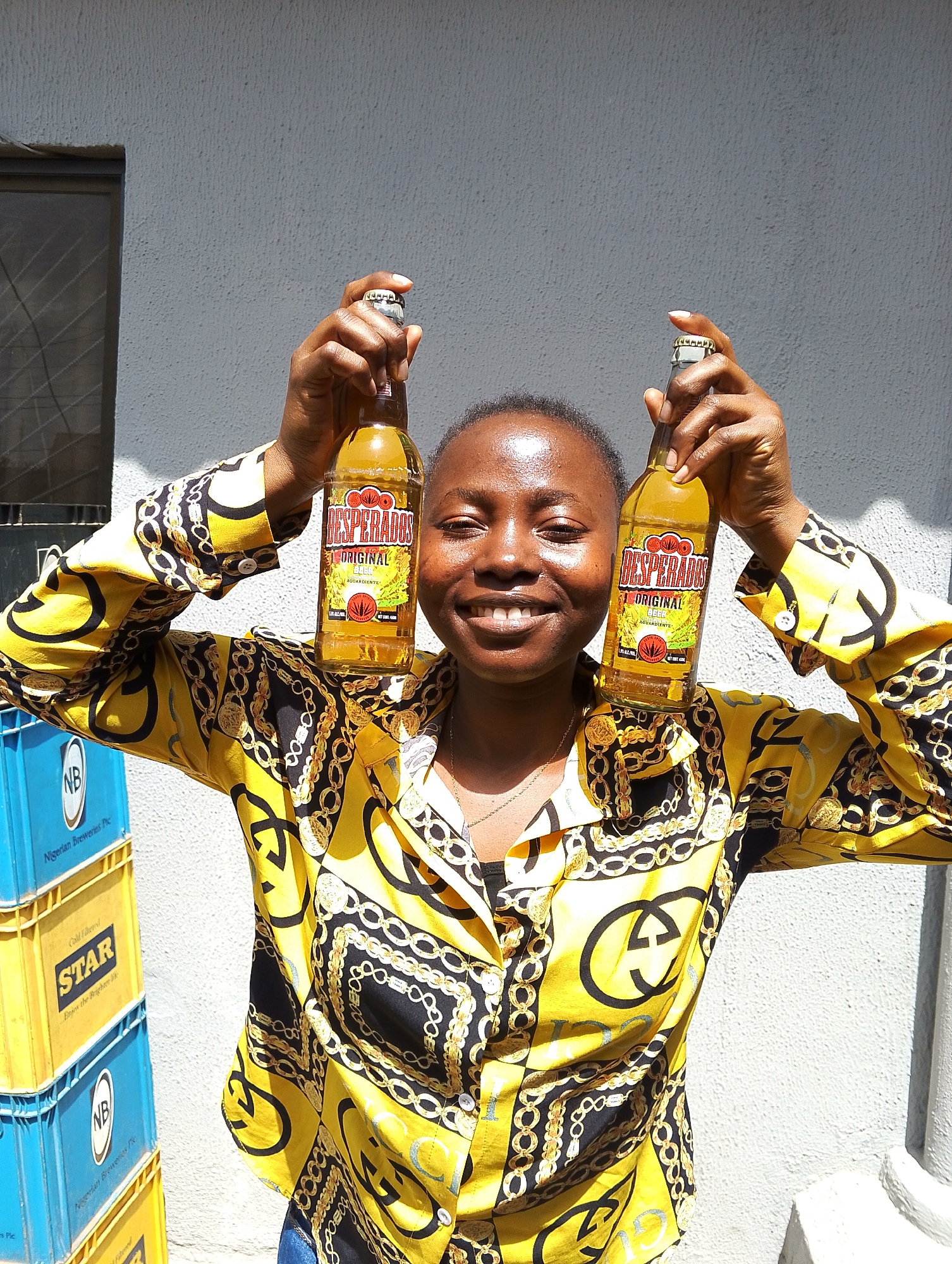A young Black woman stands outdoors, holding two bottles of Desperados Original beer above her head. She is smiling and looking towards the camera. She wears a yellow shirt with a bold black and gold chain and buckle pattern. Behind her, a stack of blue and yellow crates is visible. The crates are labeled "NB" and "STAR". The background is a textured, light gray wall with a dark window on the left side. A white pillar is visible on the right. The lighting suggests a sunny day. The presence of "Nigerian Breweries Plc" on the crates indicates the location is likely within or near a facility associated with this company in Jos, Nigeria.